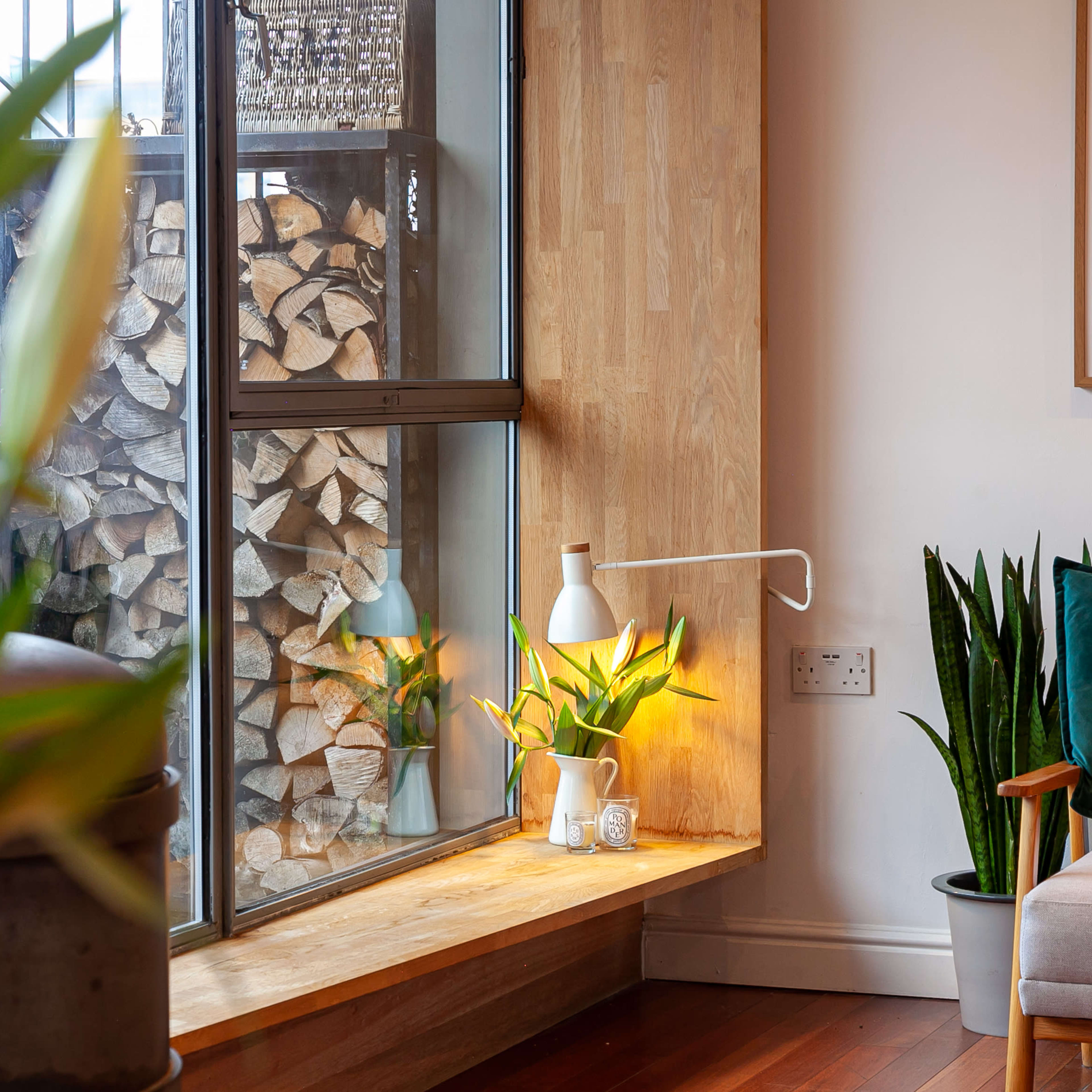 A well-lit corner of a living room features a wooden window sill with plants, a lamp, and a stack of logs visible through the glass.