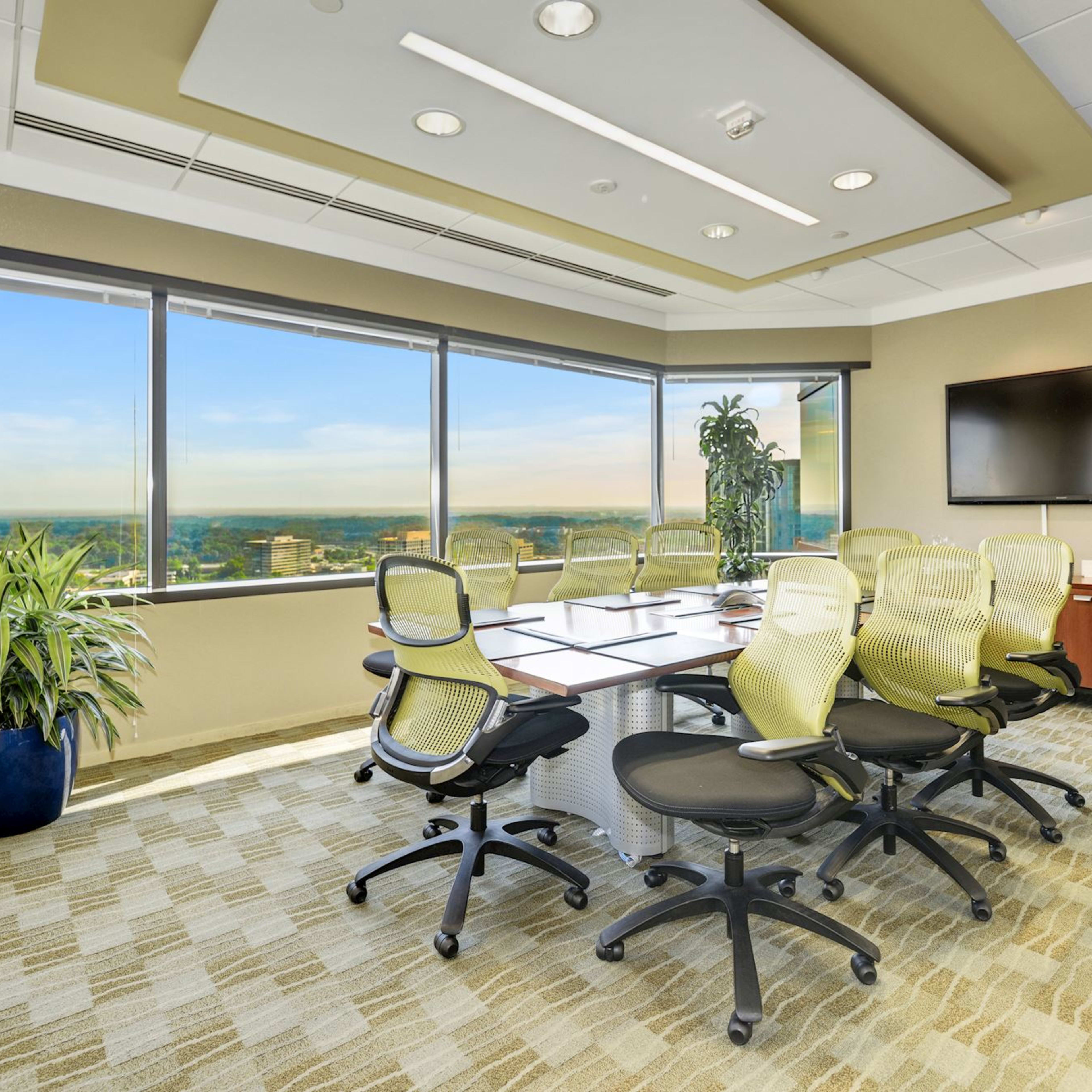 A modern conference room features a large table surrounded by chairs, with floor-to-ceiling windows offering a view of the city.