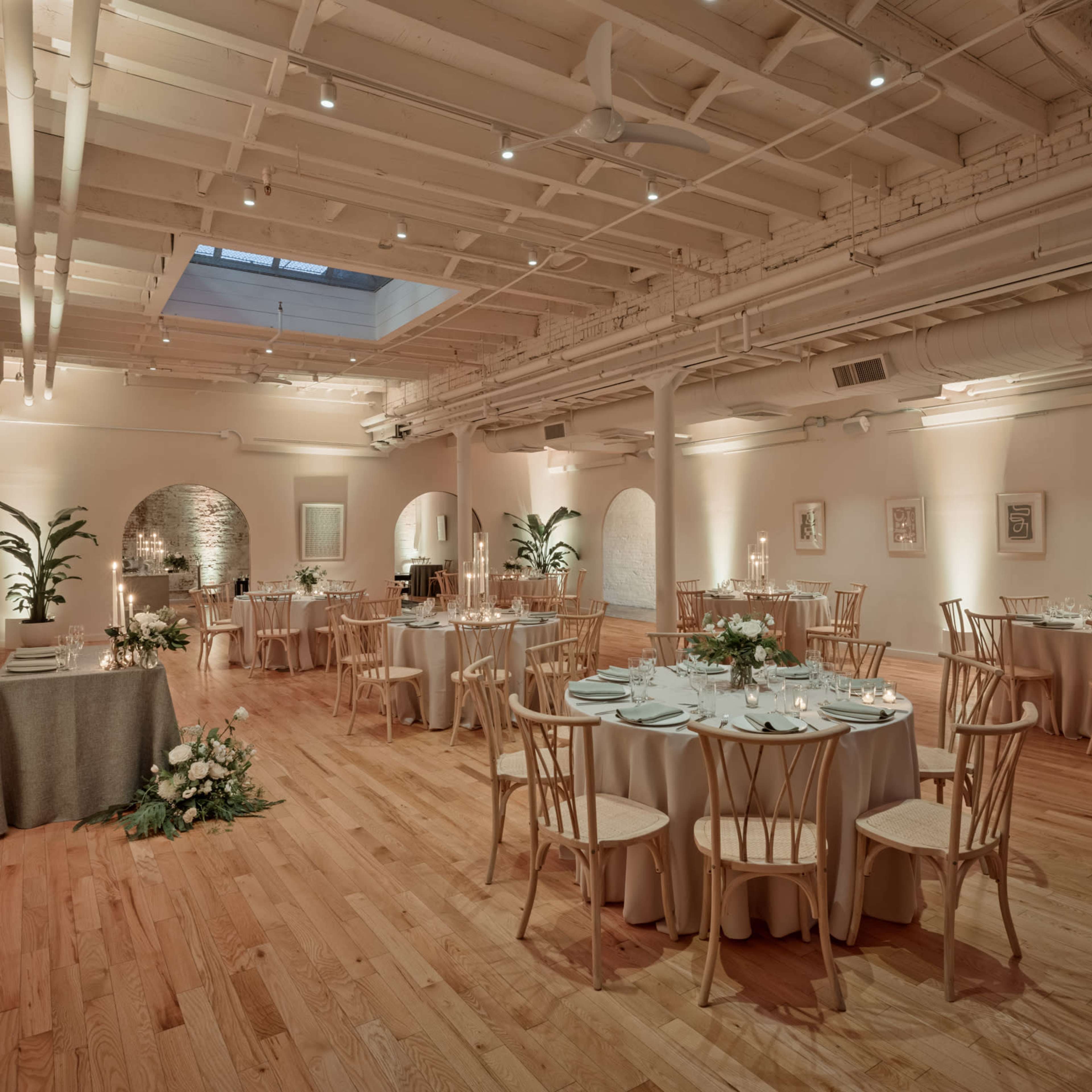 A spacious, elegantly arranged event venue with round tables set for dining, surrounded by light wood flooring and soft lighting.
