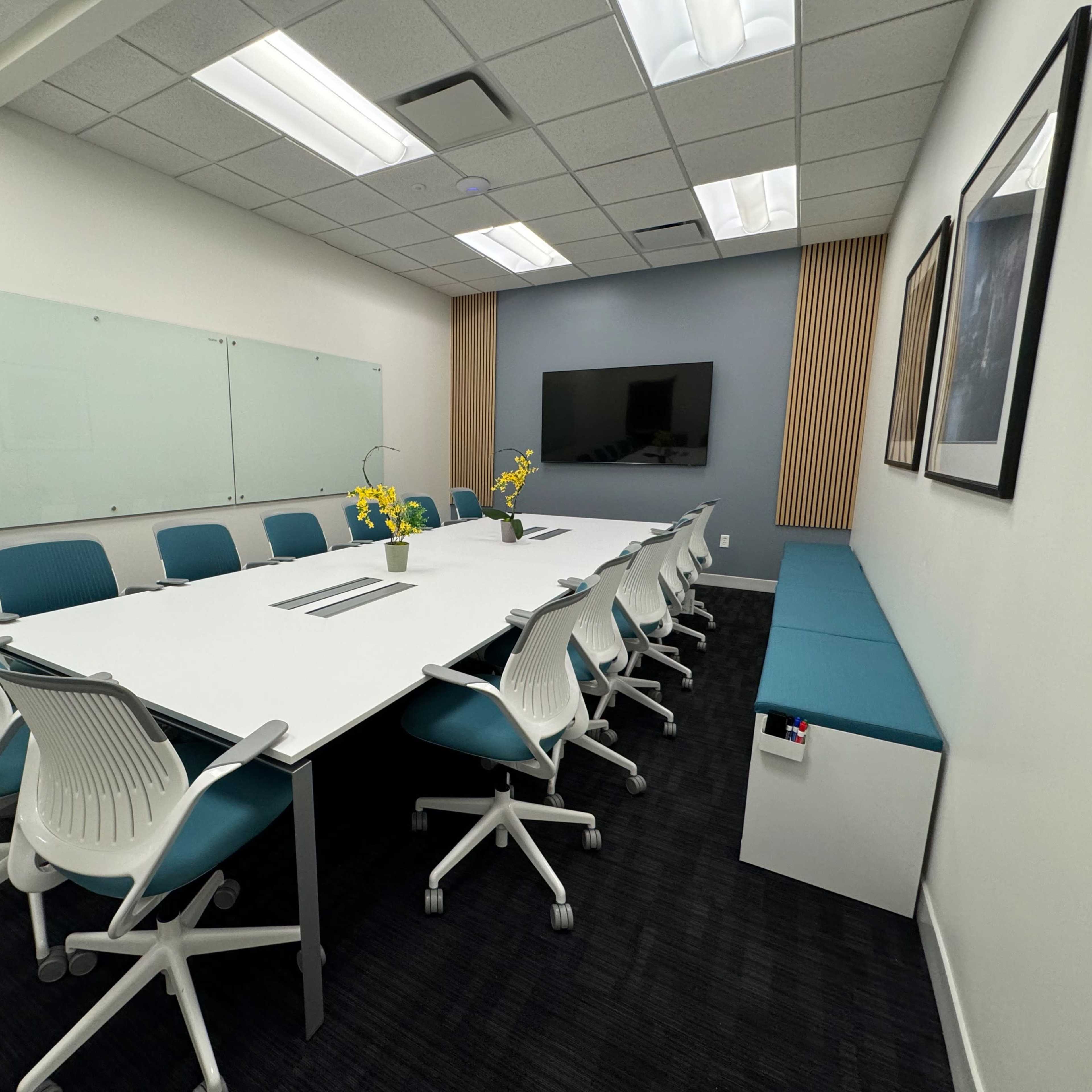 A modern conference room featuring a large white table surrounded by blue and white chairs, with a wall-mounted television and a whiteboard.