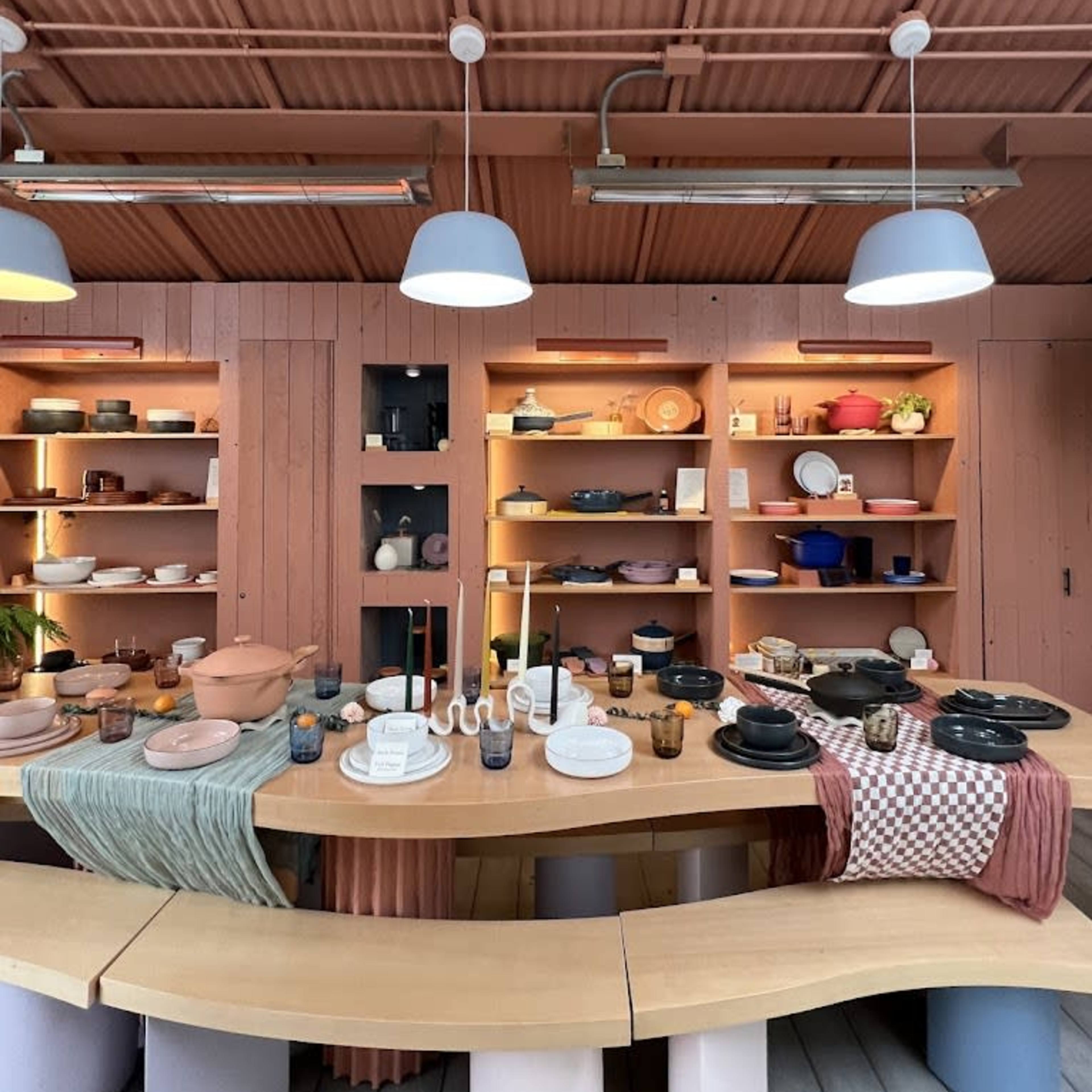 A modern kitchen interior with a curved wooden table surrounded by various dishes, cookware, and decorative items displayed on shelves.