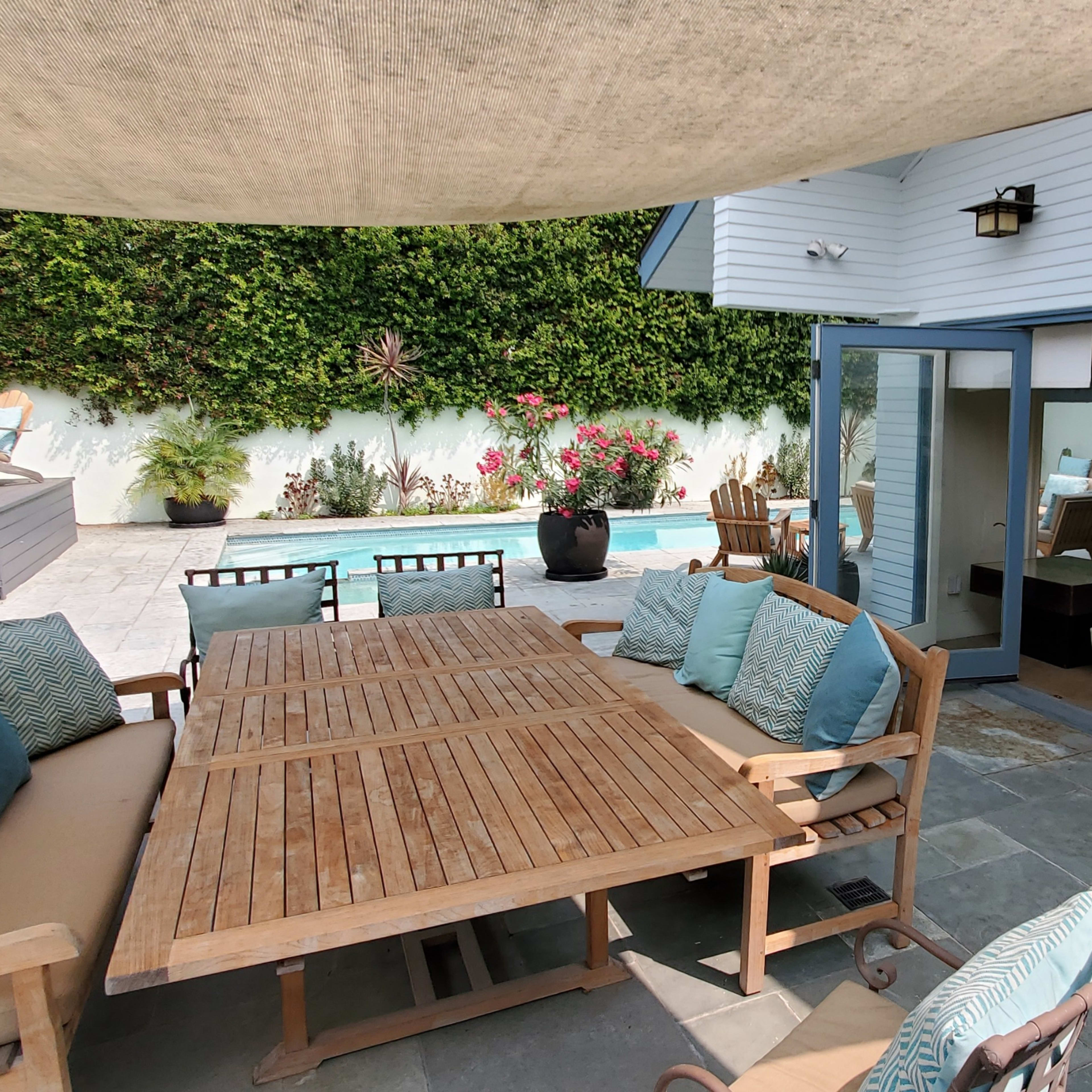 A patio area features a wooden dining table surrounded by chairs, with a pool visible in the background and a lounge area inside the house.