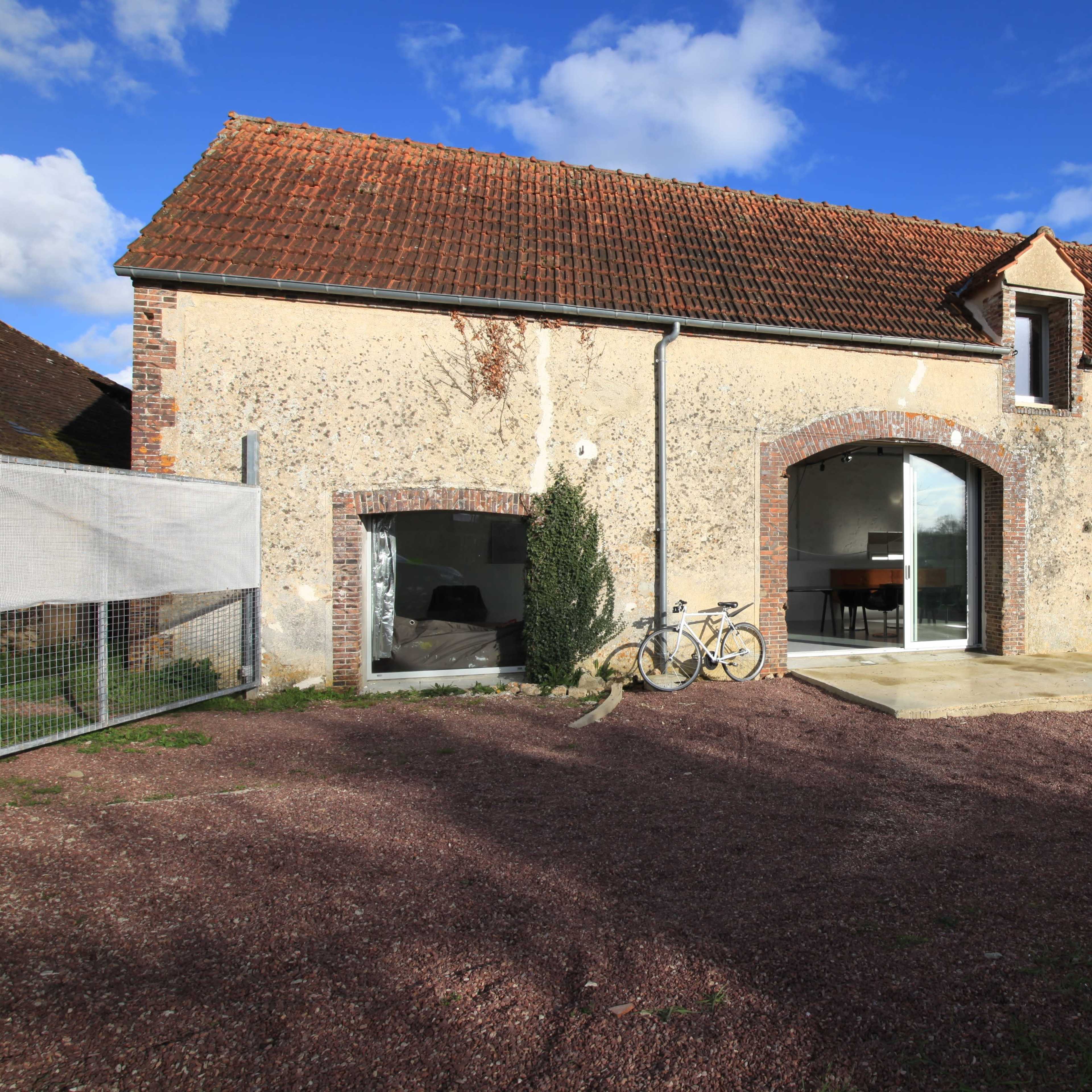 A stone house with a red-tiled roof features large windows and a small patio area, set against a blue sky with scattered clouds.
