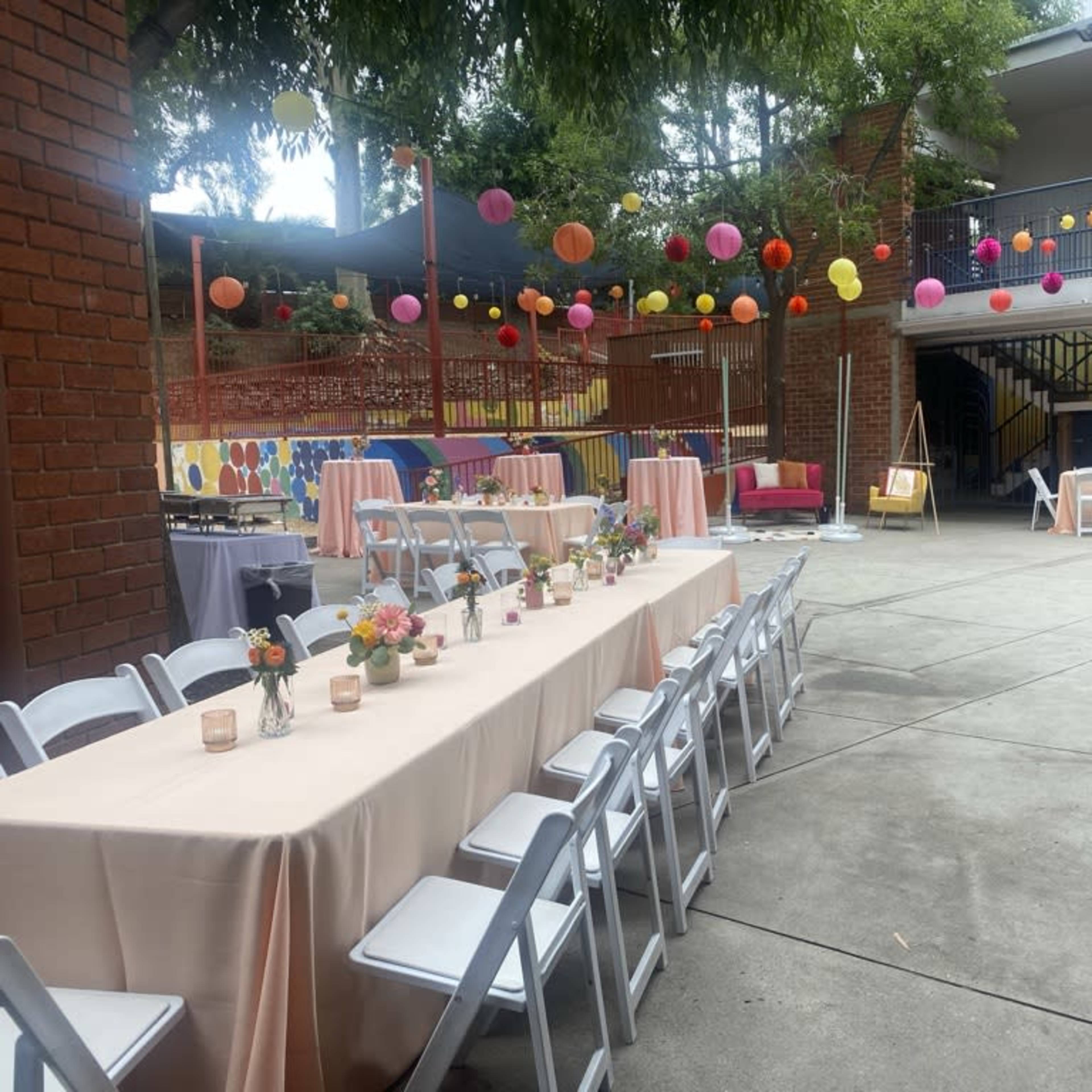 A courtyard is set up for a celebration, featuring decorated tables, colorful paper lanterns hanging above, and vibrant party decorations.