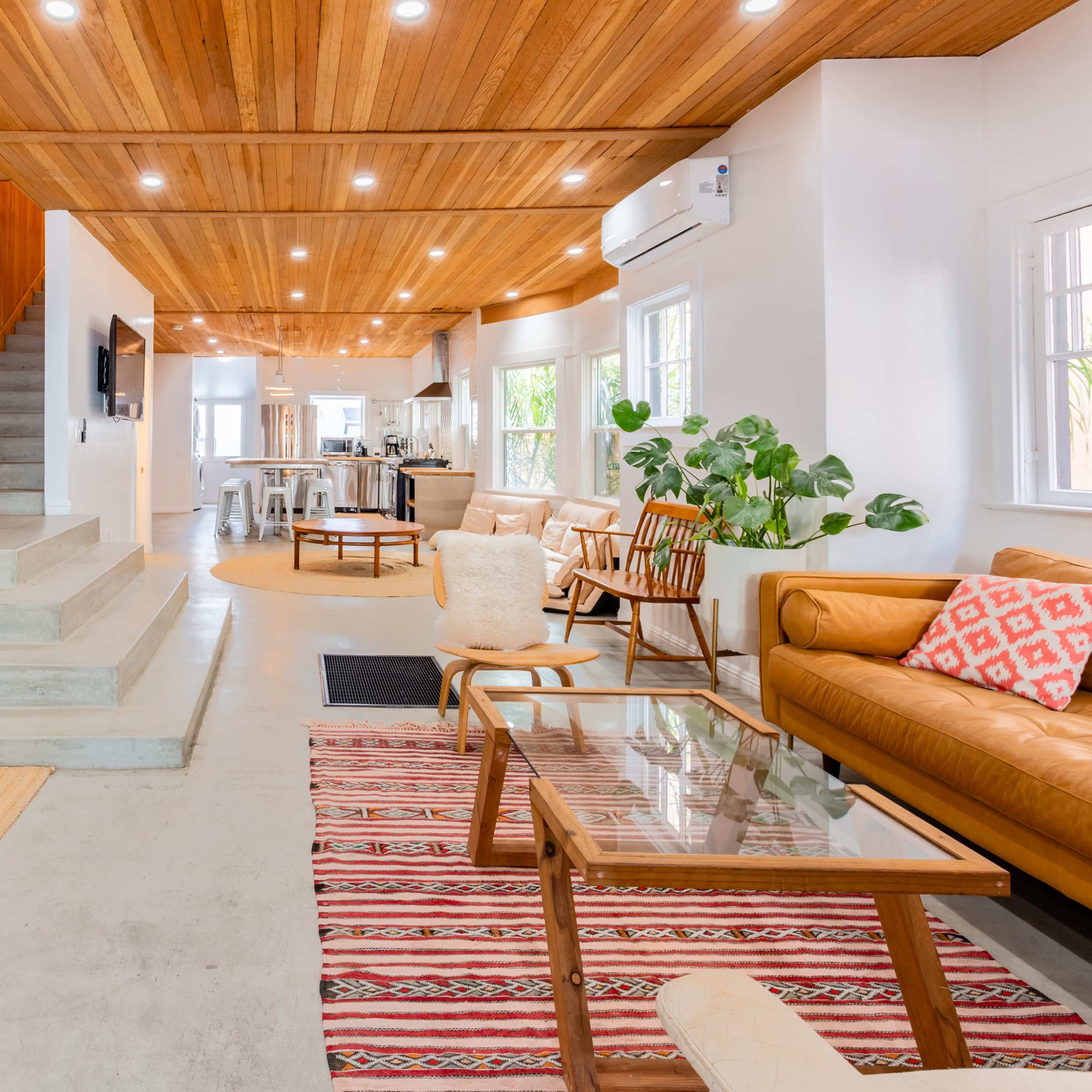 A modern living room with wooden ceilings, a staircase leading to an upper level, and a mix of seating areas, including a leather sofa and a glass coffee table.