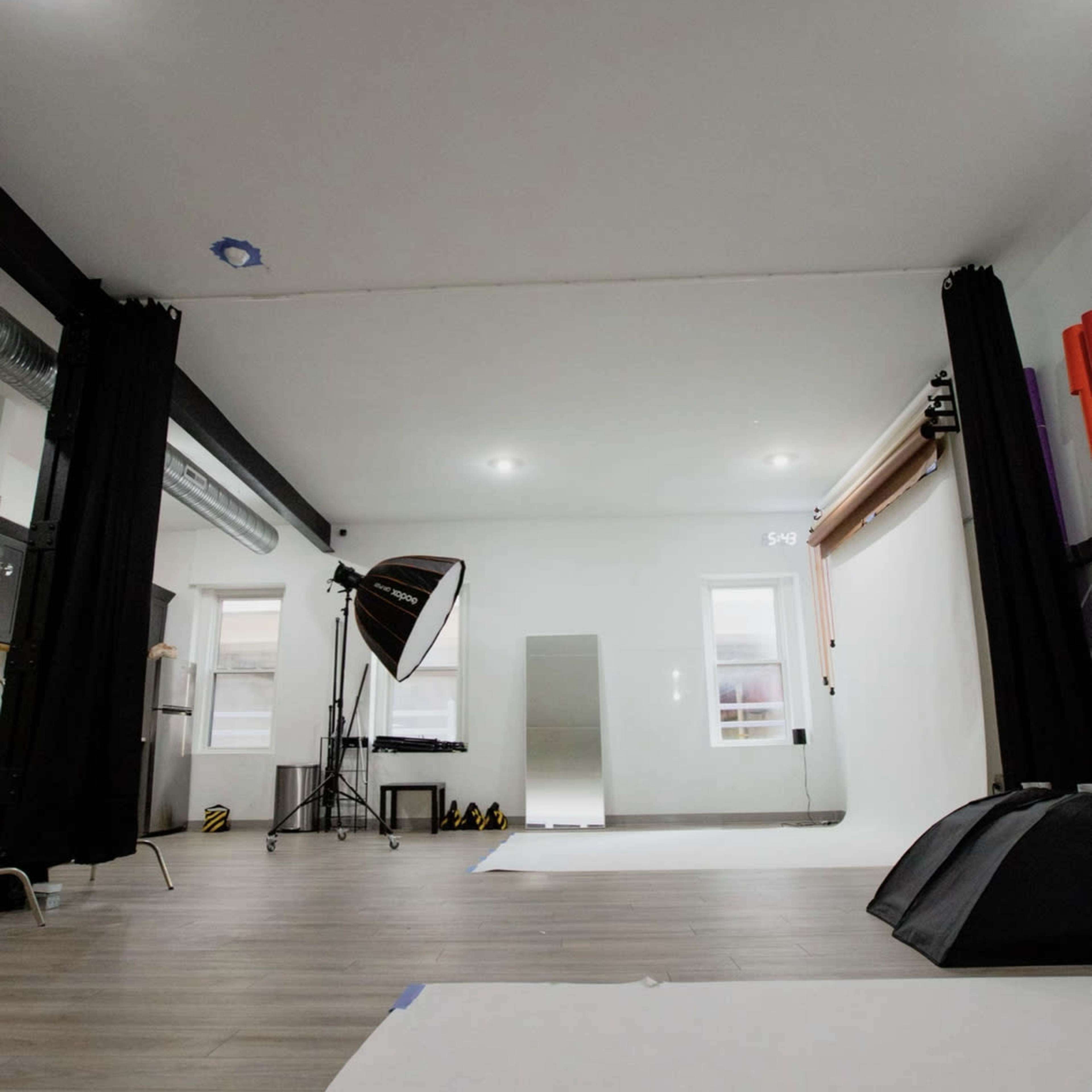 A photography studio features a large mirror, backdrop, softbox light, and plain wooden flooring.