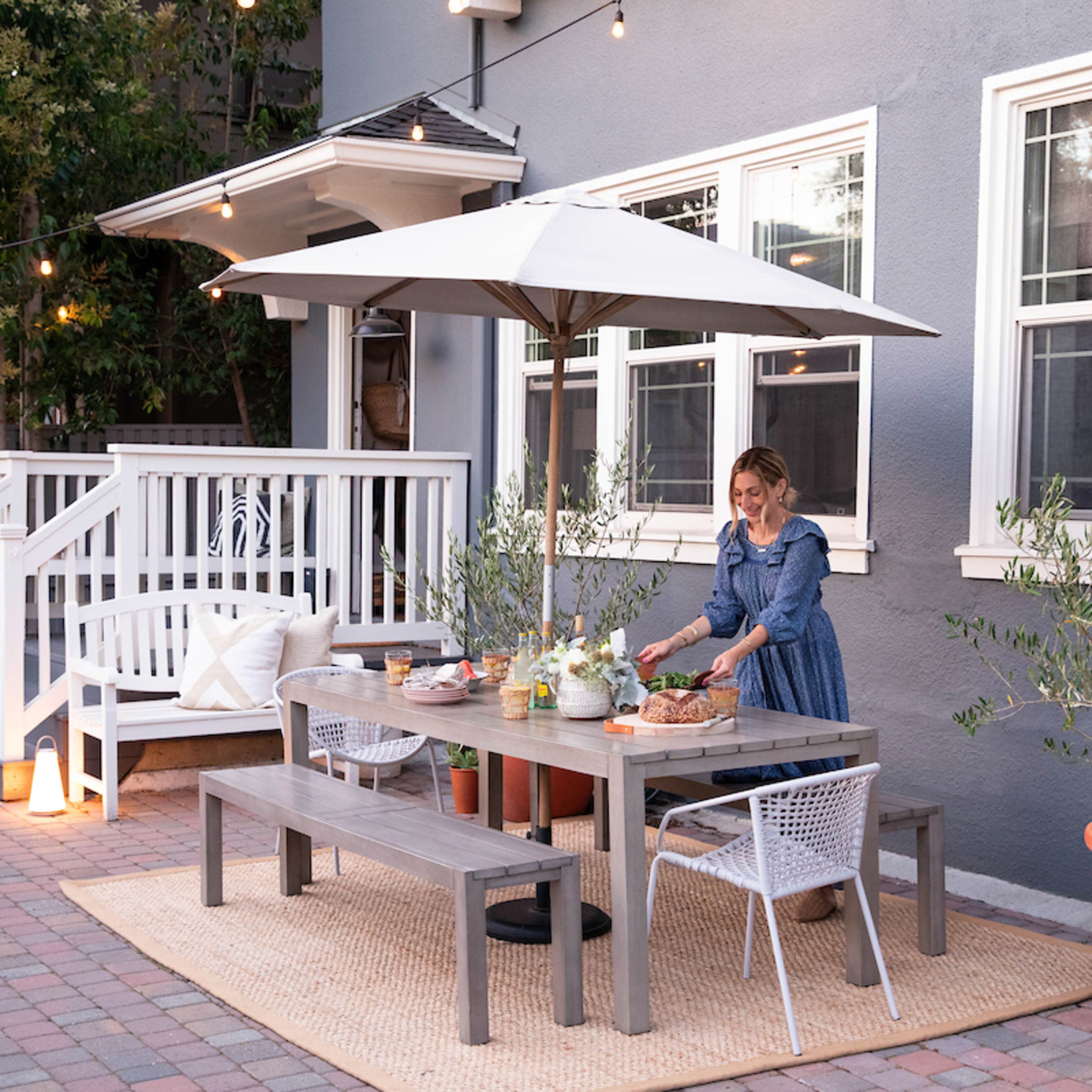 A woman sets the table on a patio with a large umbrella, surrounded by potted plants and string lights.