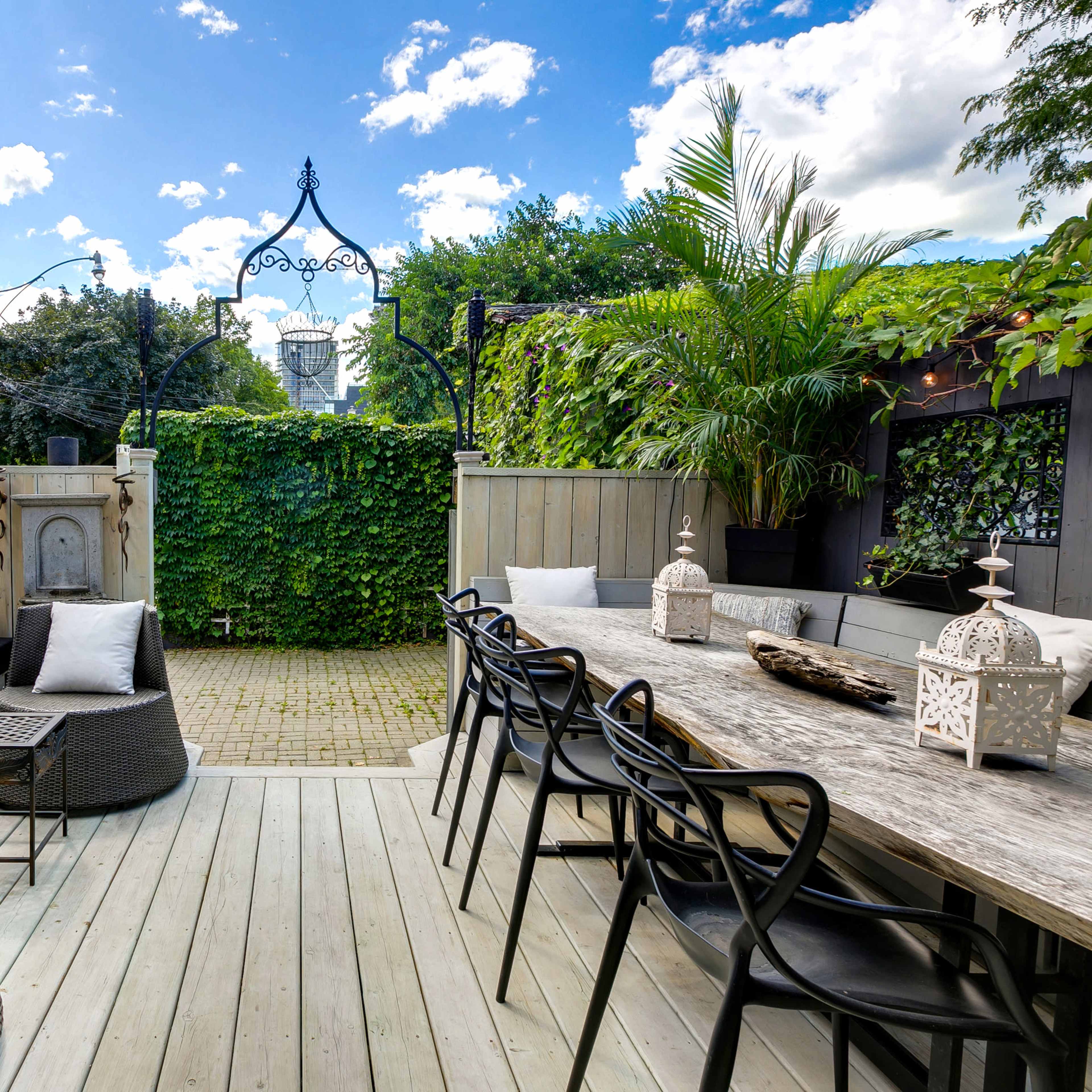 A landscaped outdoor deck area featuring a seating arrangement with a black couch, a dining table, and lush greenery surrounding the space.