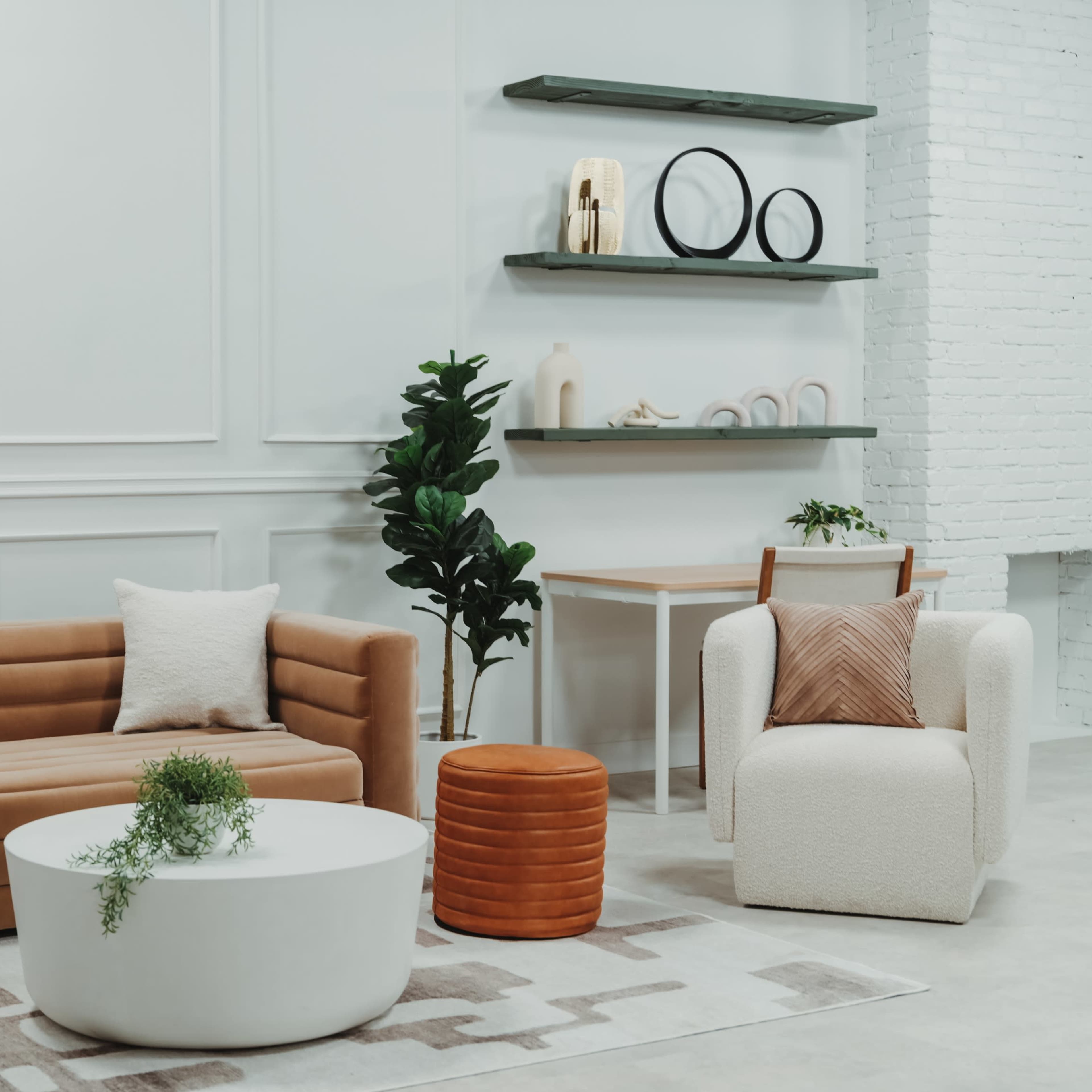 A modern living room features a brown sofa, a white circular coffee table, and a light-colored armchair, surrounded by minimalist decor and greenery.