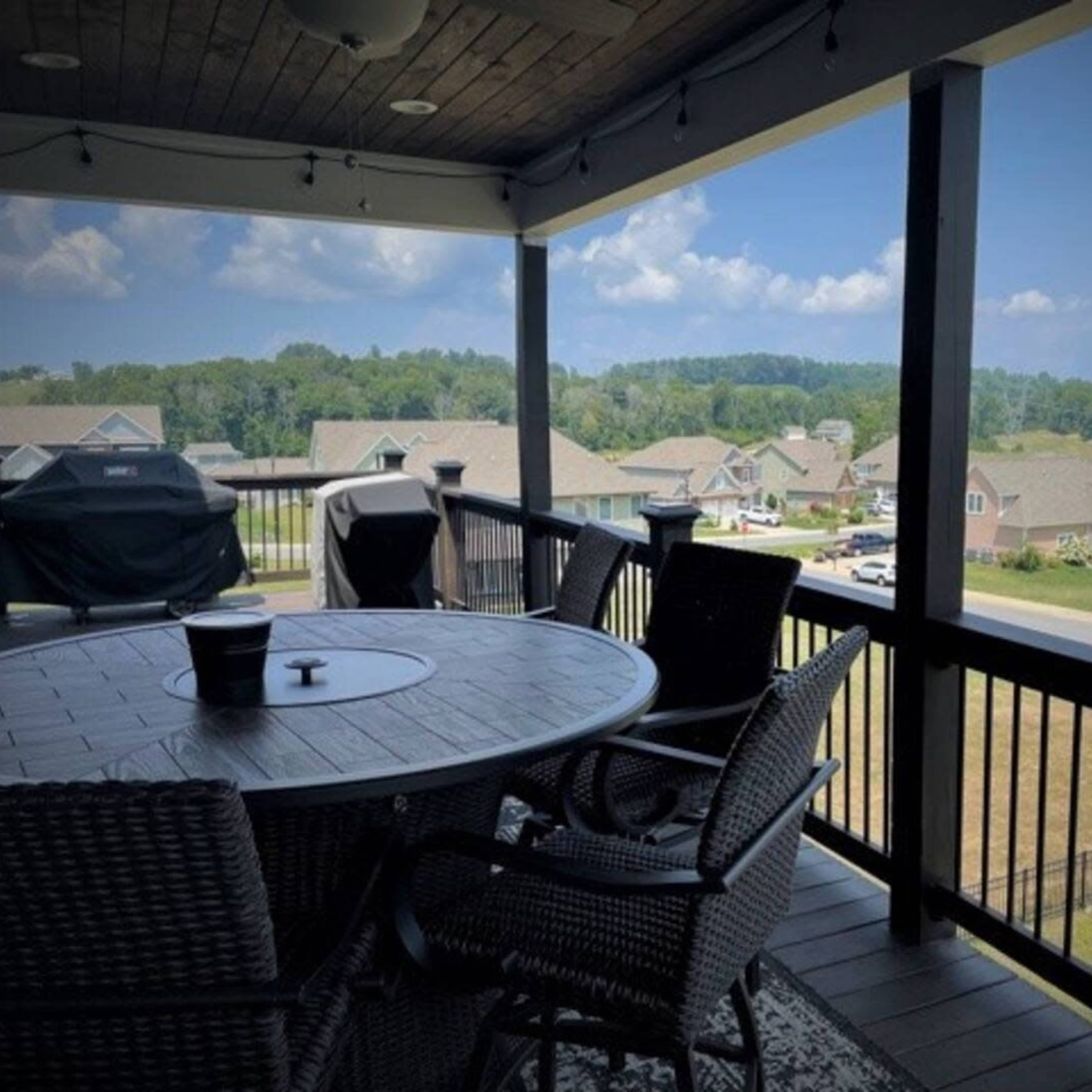 A covered balcony features a round table and chairs with a view of suburban homes and greenery in the background.