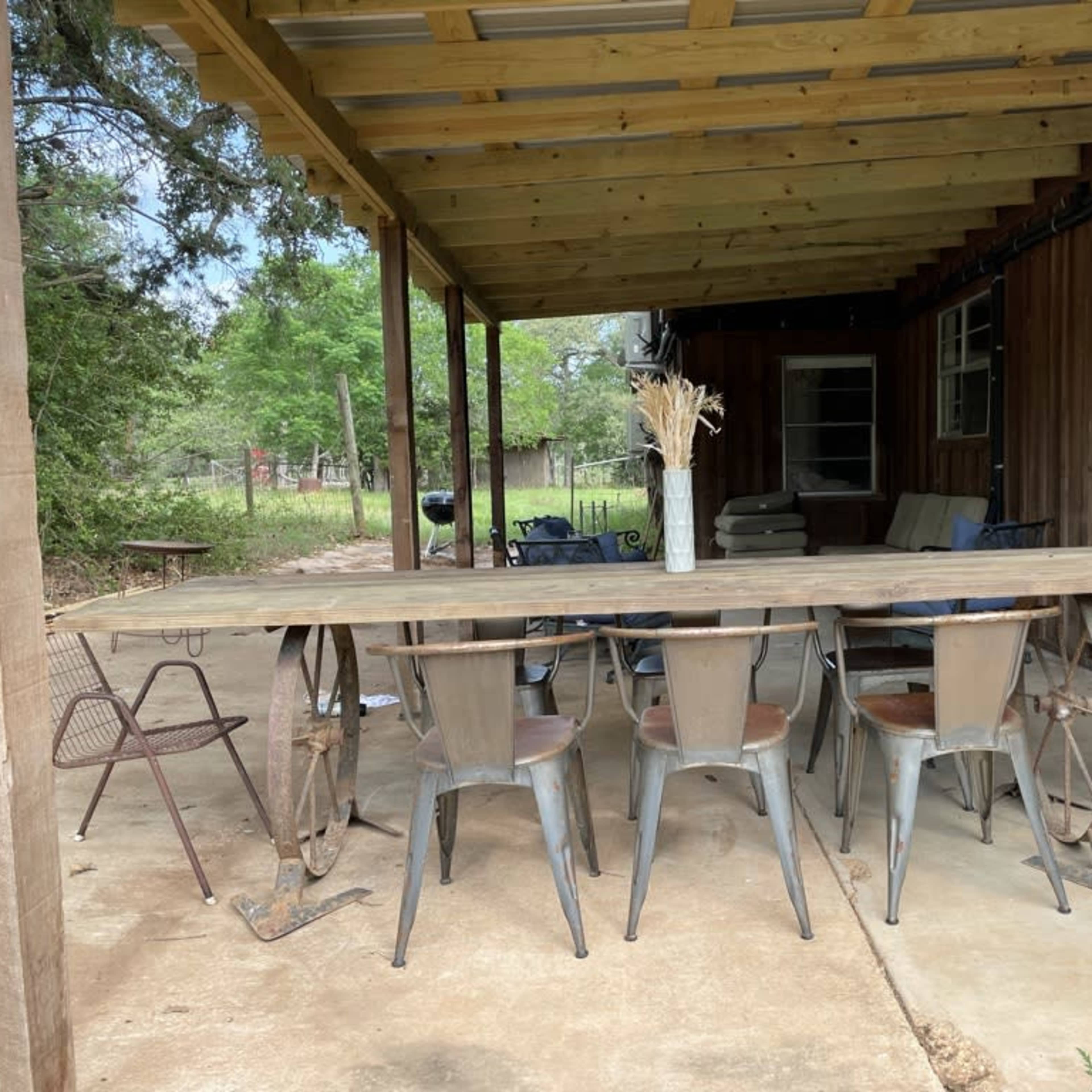 A long wooden table is set under a covered porch surrounded by metal chairs in a rustic outdoor setting.