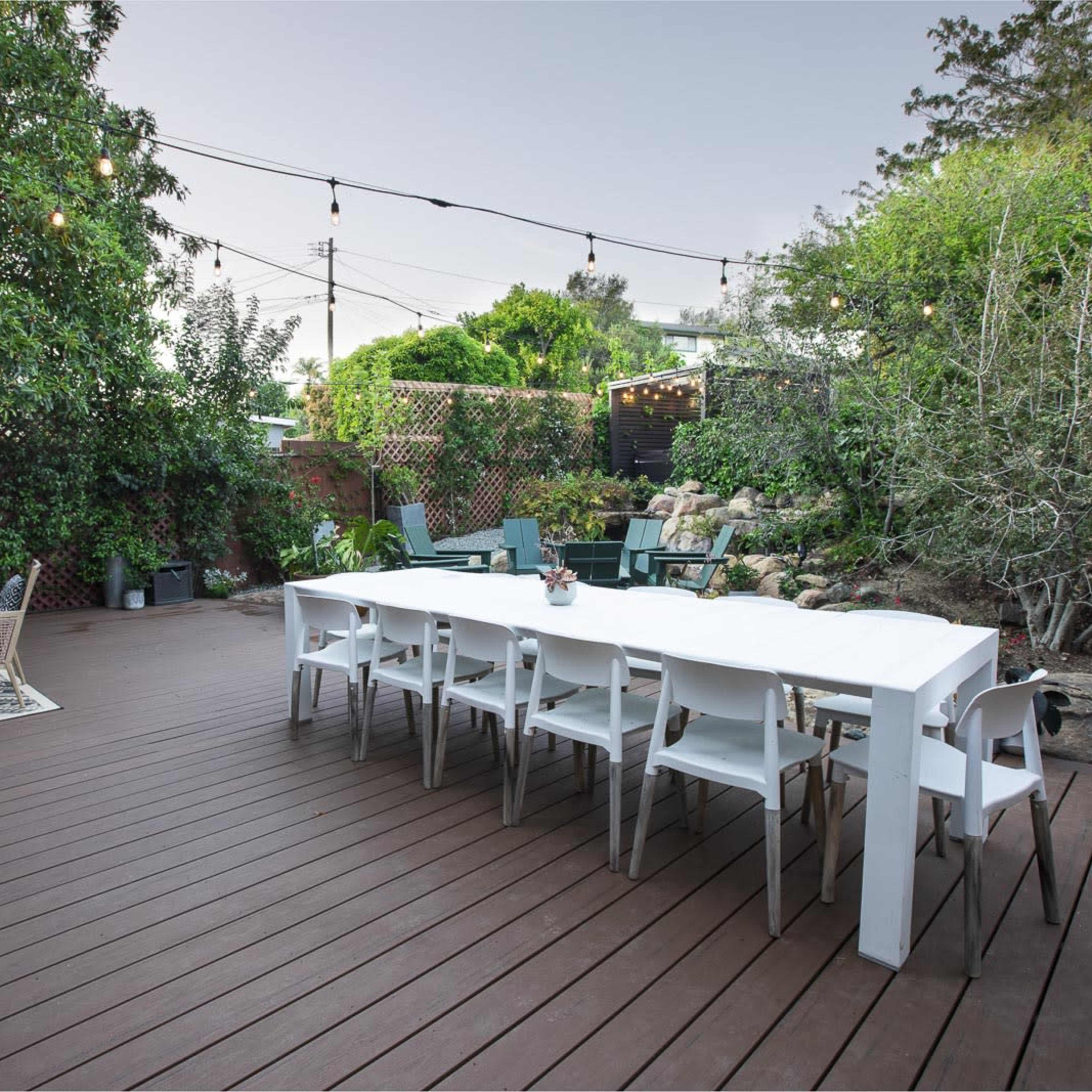 A spacious outdoor patio with a long white dining table, surrounded by chairs, and adorned with string lights and greenery.