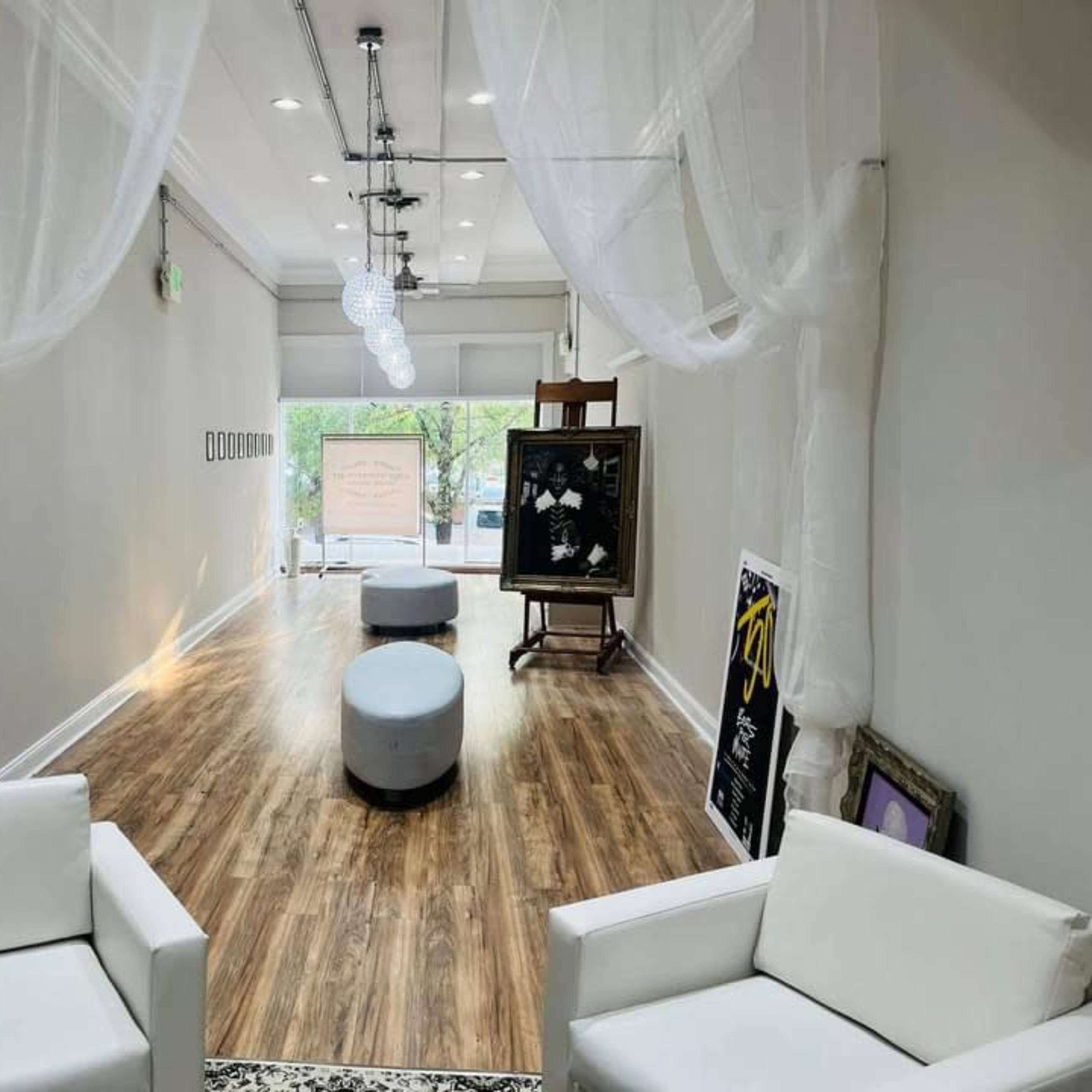 A spacious hallway with white walls, wooden flooring, two white chairs, and round gray ottomans, illuminated by hanging light fixtures and draped fabric.