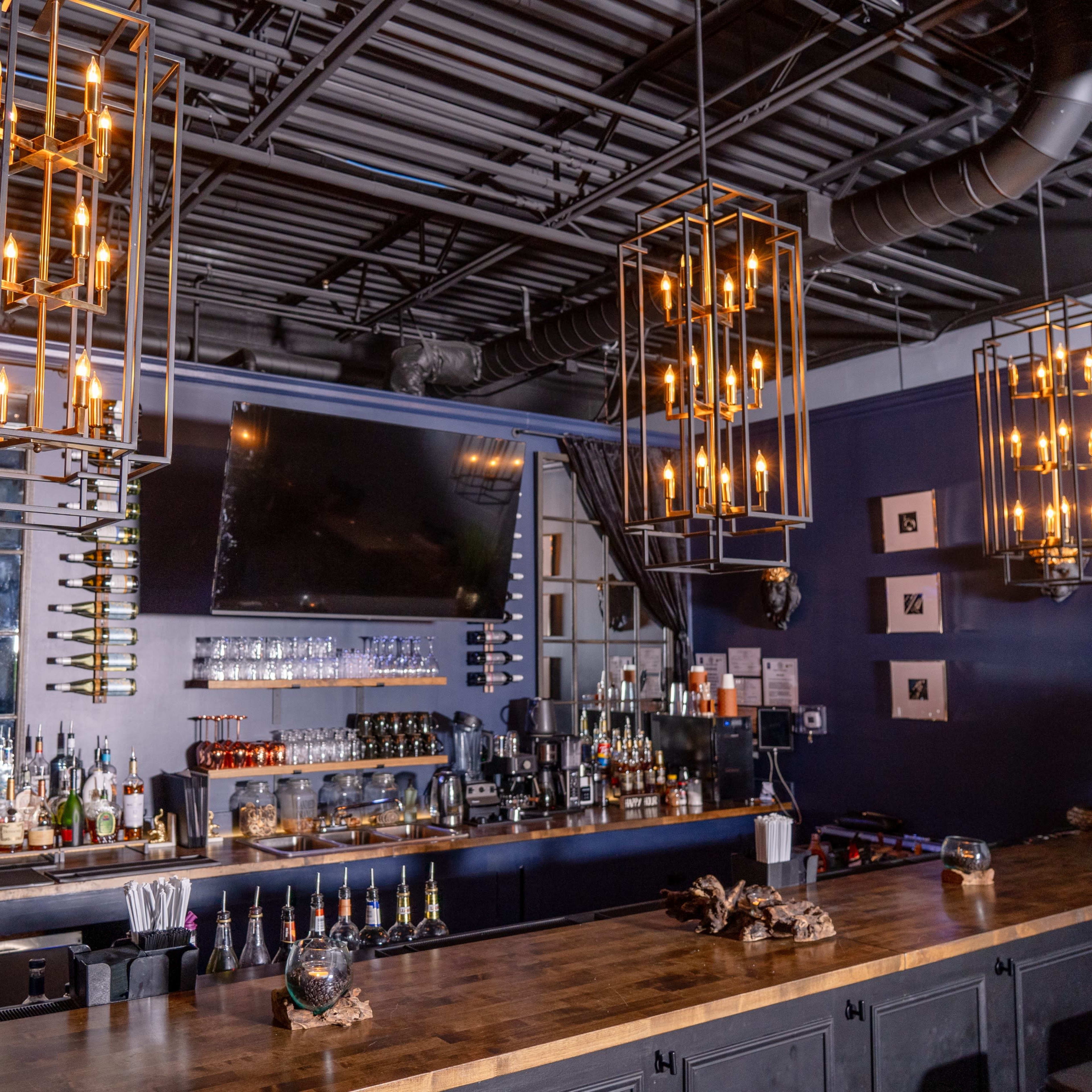 A modern bar with a long wooden counter, overhead pendant lights, a selection of bottles on the shelves, and a large TV screen mounted on the wall behind it.