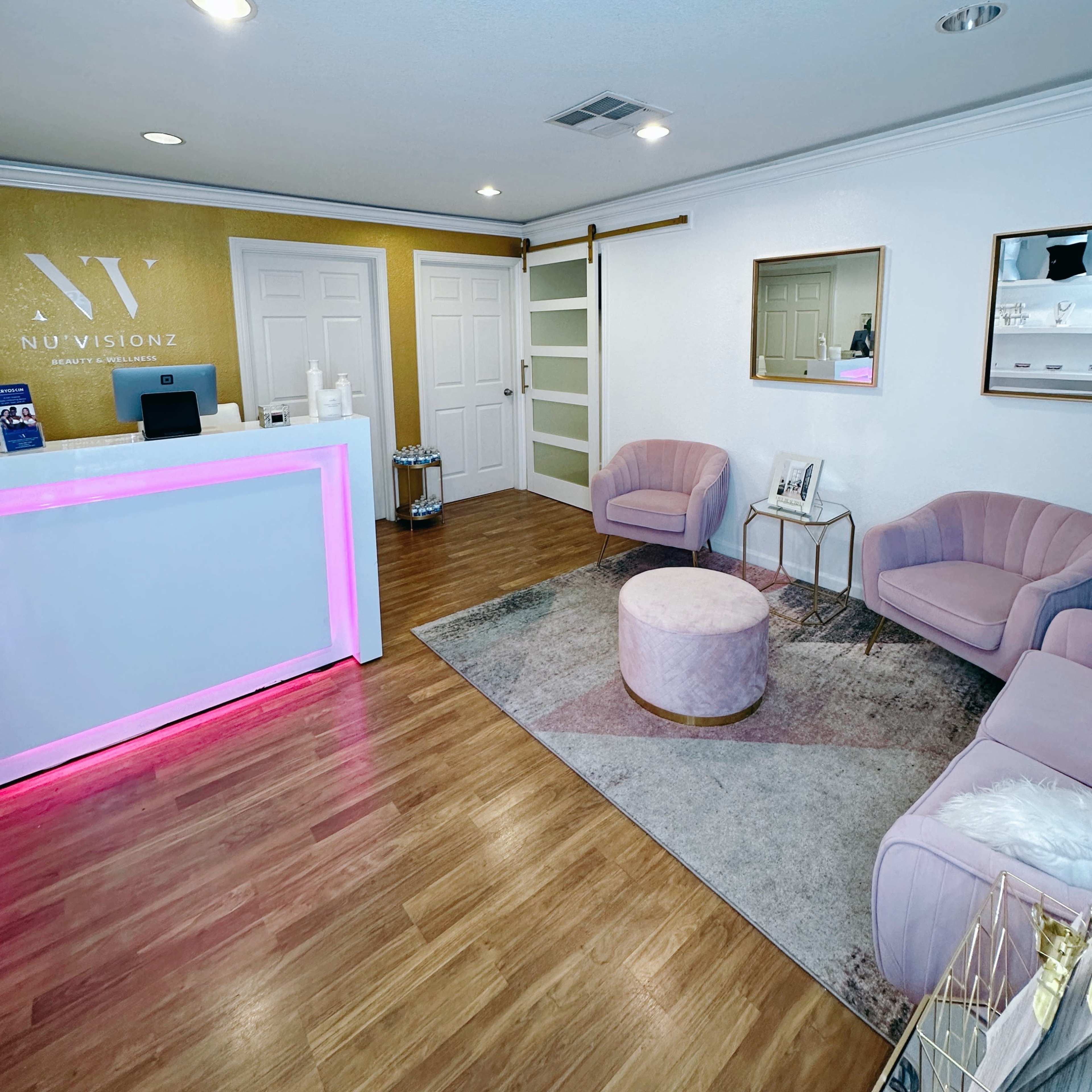 A modern reception area with a white front desk illuminated in pink, two pink chairs, a round ottoman, and a decorative rug.