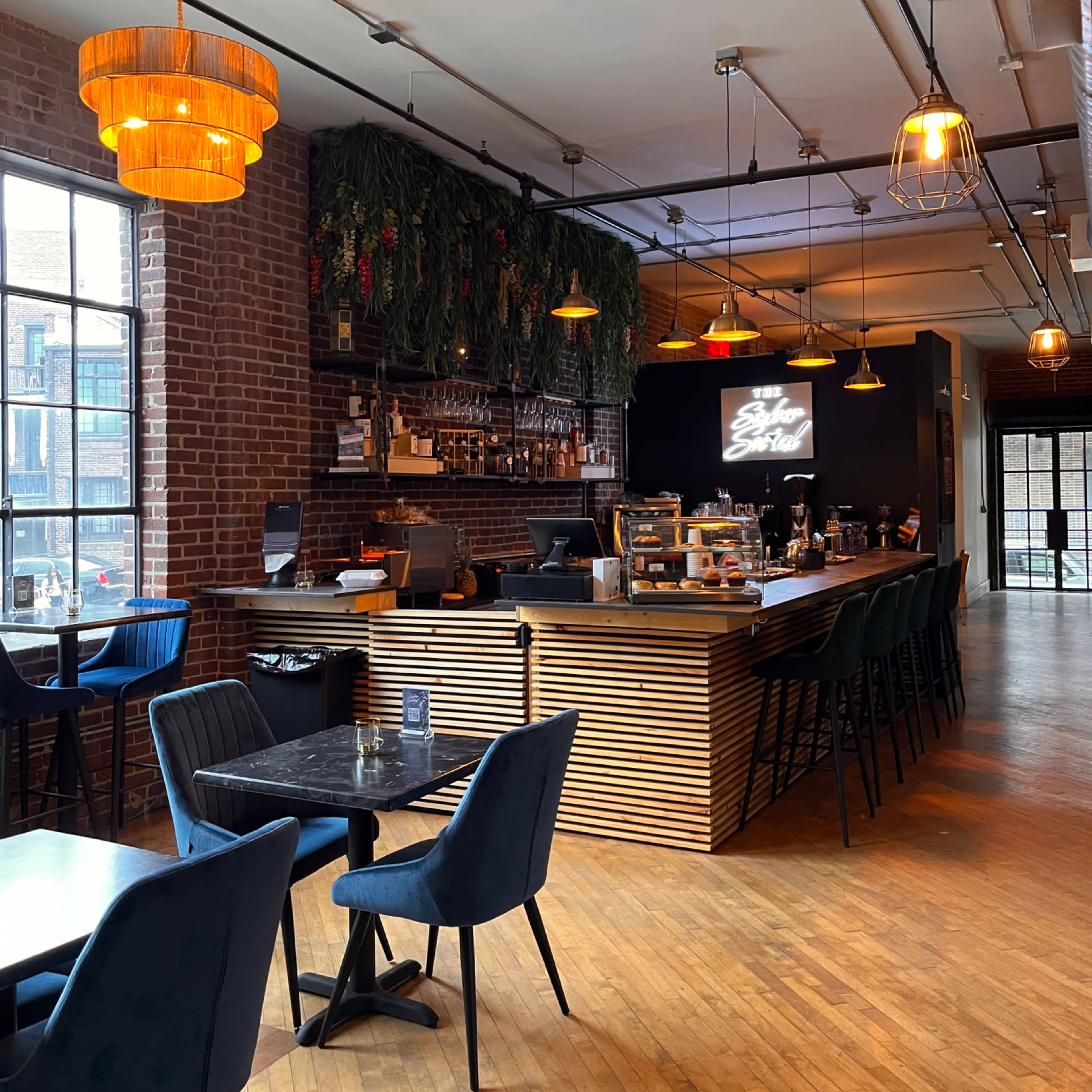 A modern café interior with wooden floors, a bar area adorned with hanging lights, and a mix of seating options including dark chairs and a black table.