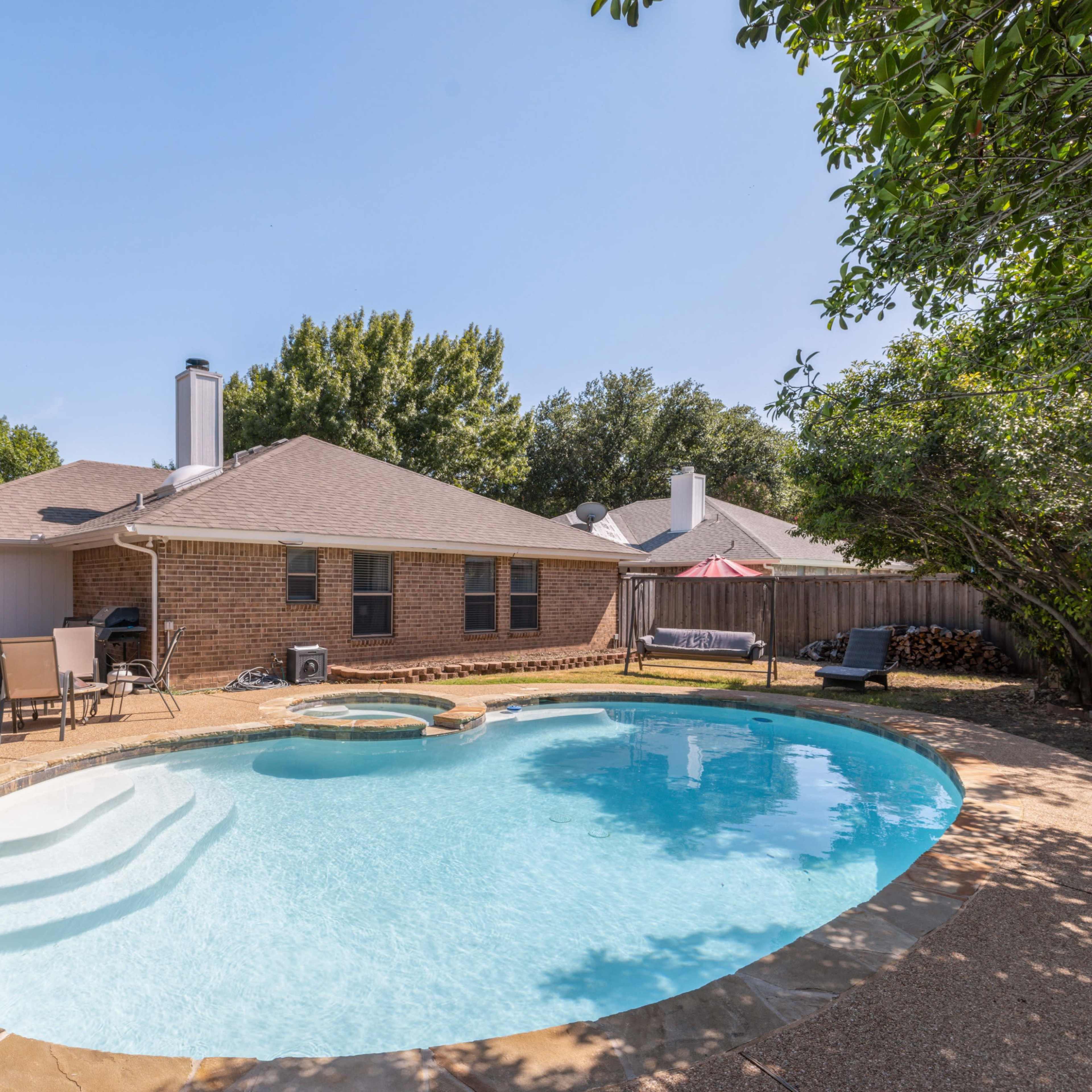 A backyard with a curved swimming pool surrounded by light brown stone, a small hot tub, and a brick house in the background.