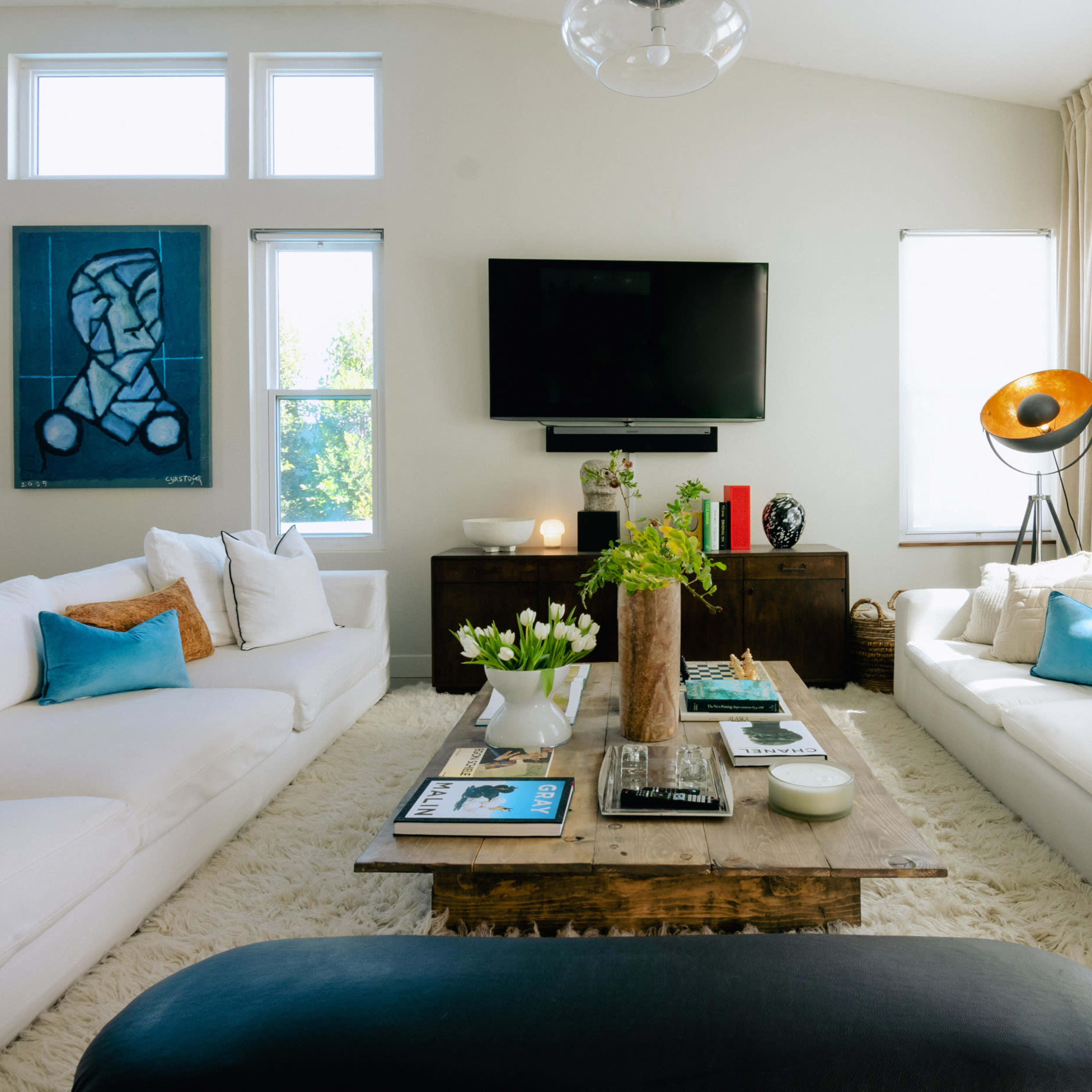 A spacious living room features white sofas, a wooden coffee table, a large TV on the wall, and framed artwork beside large windows.