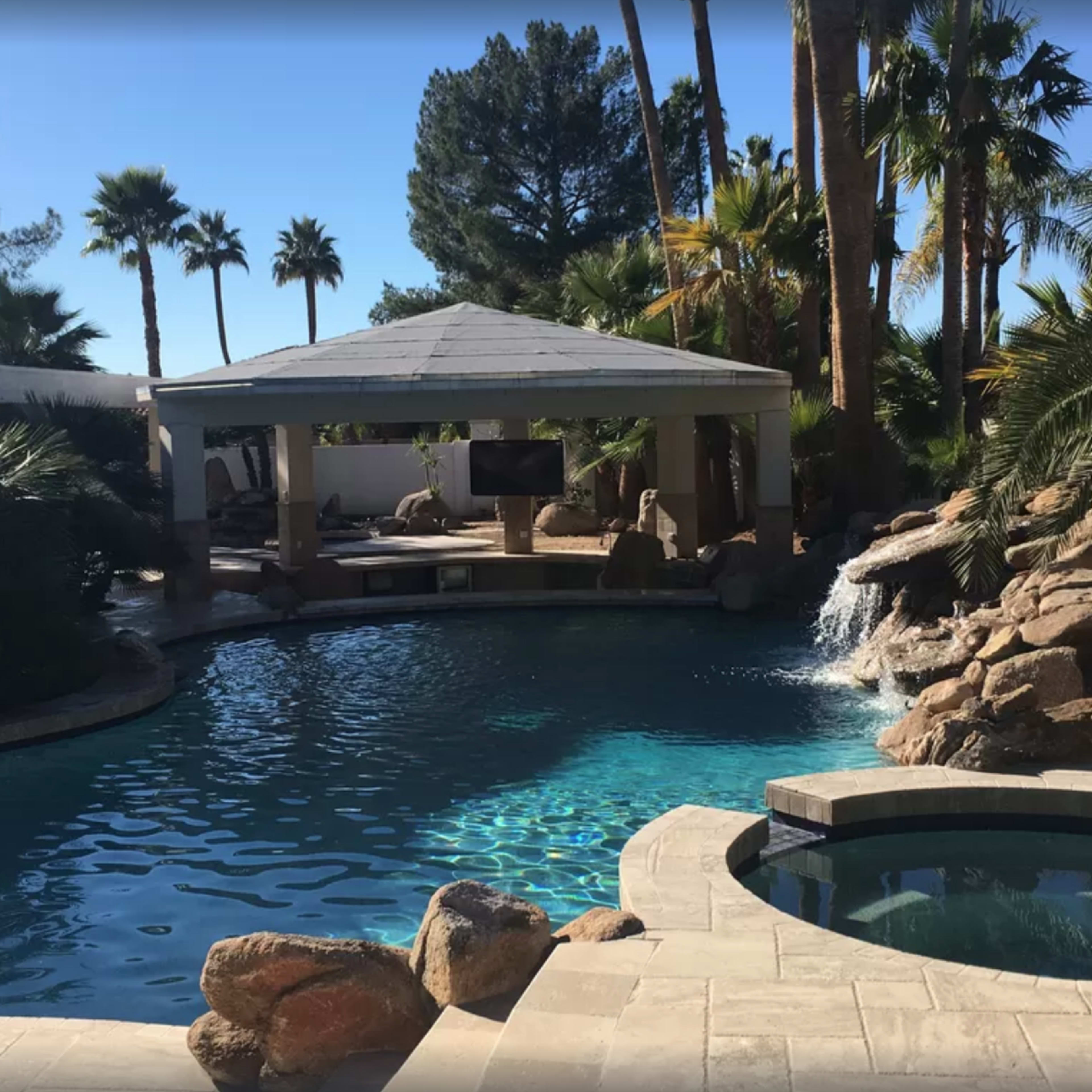 A large swimming pool surrounded by palm trees, a gazebo, and a rocky waterfall feature.