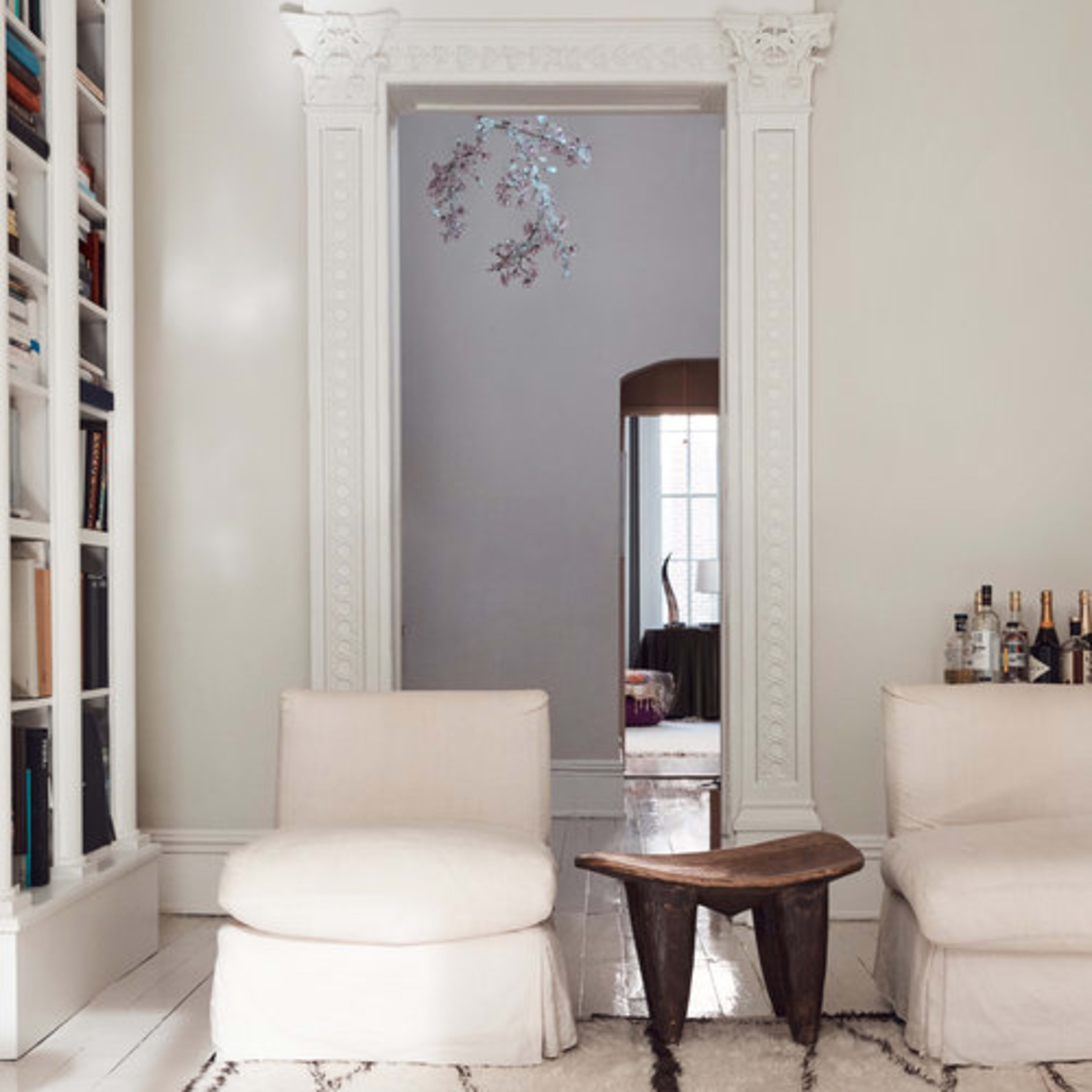 A minimalist living room features two white chairs, a wooden table, and a doorway with ornate trim leading to another room.