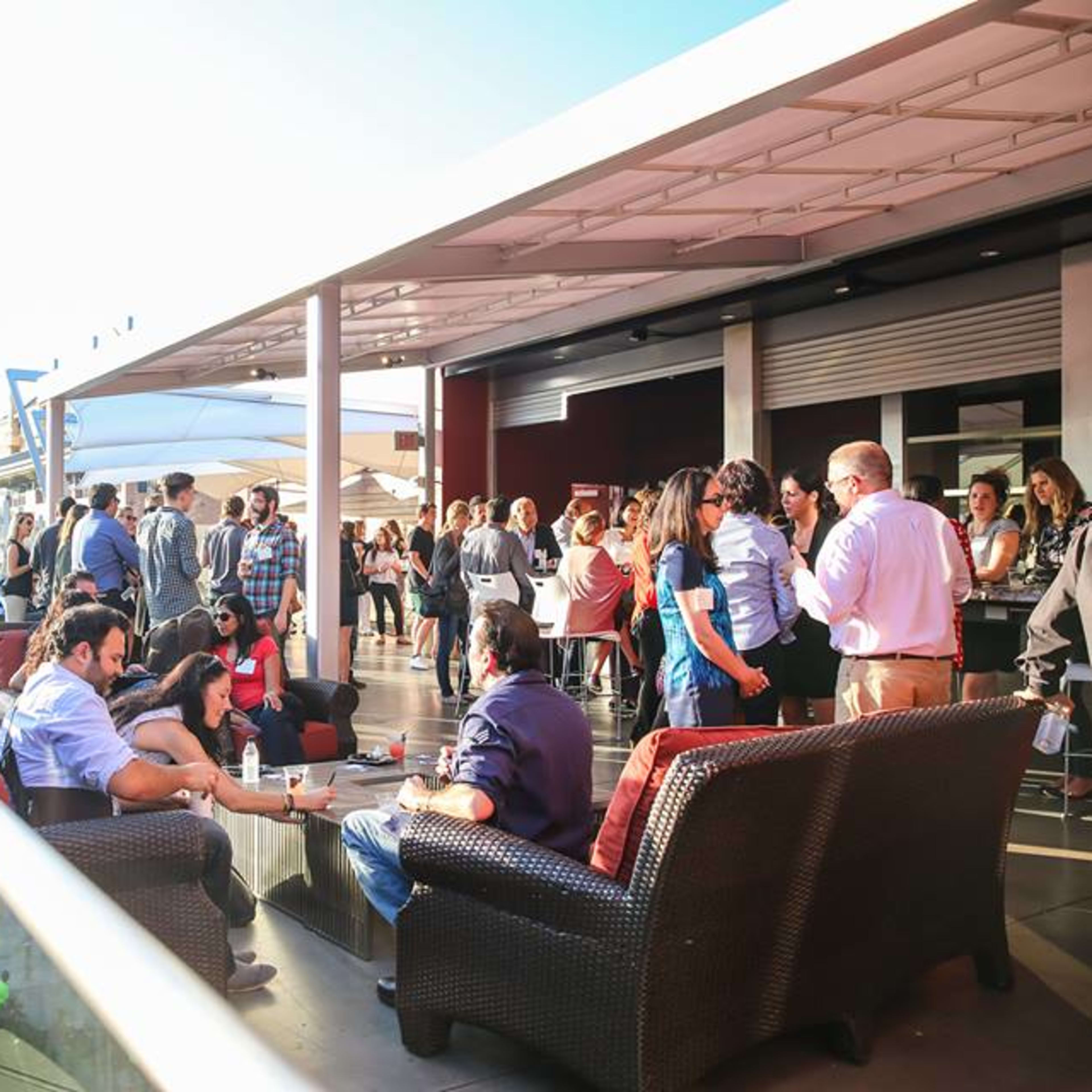 A group of people socializes on a rooftop terrace, enjoying a sunny day with a view of a pedestrian street below.