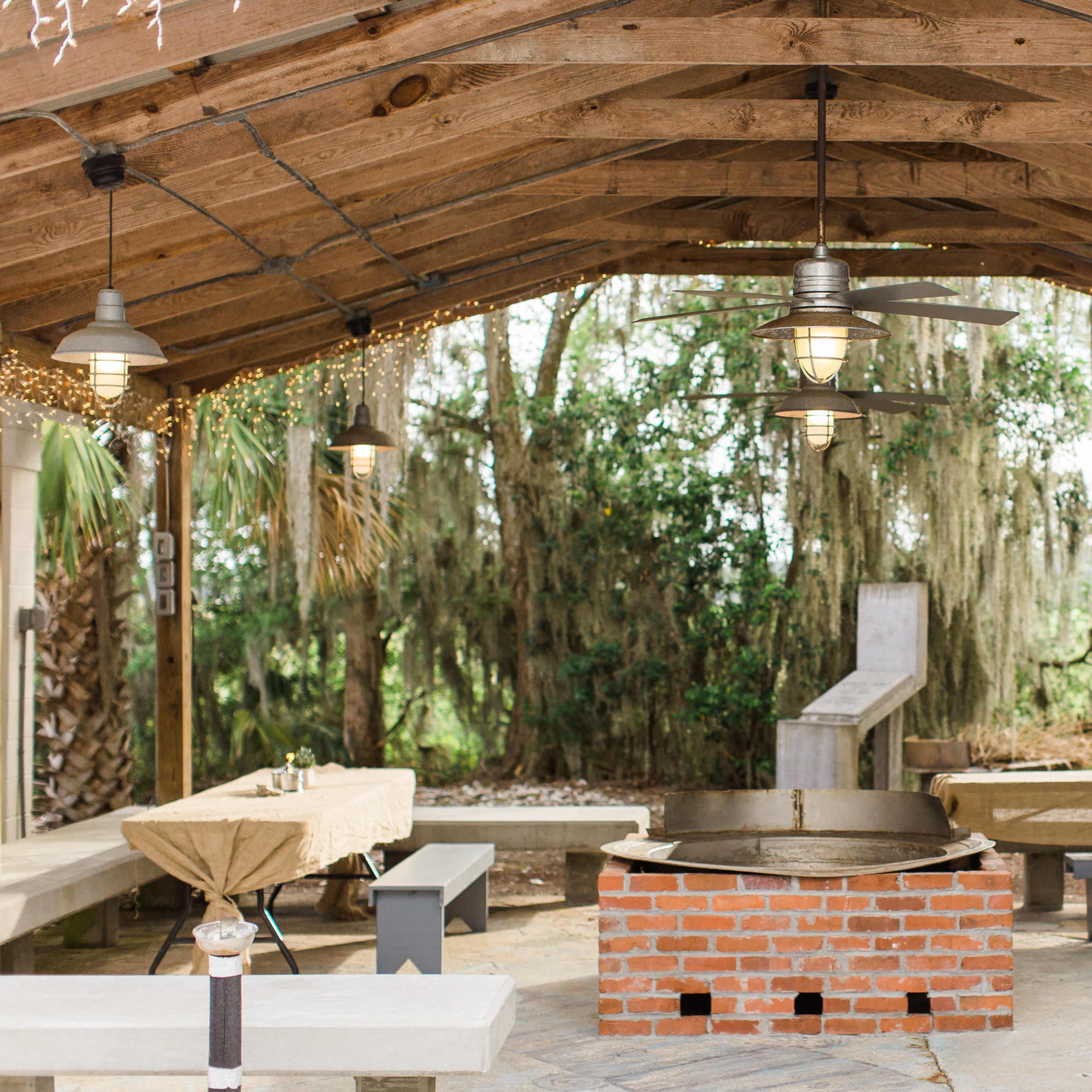 A covered outdoor gathering space with a brick fire pit in the center, surrounded by several tables and benches.