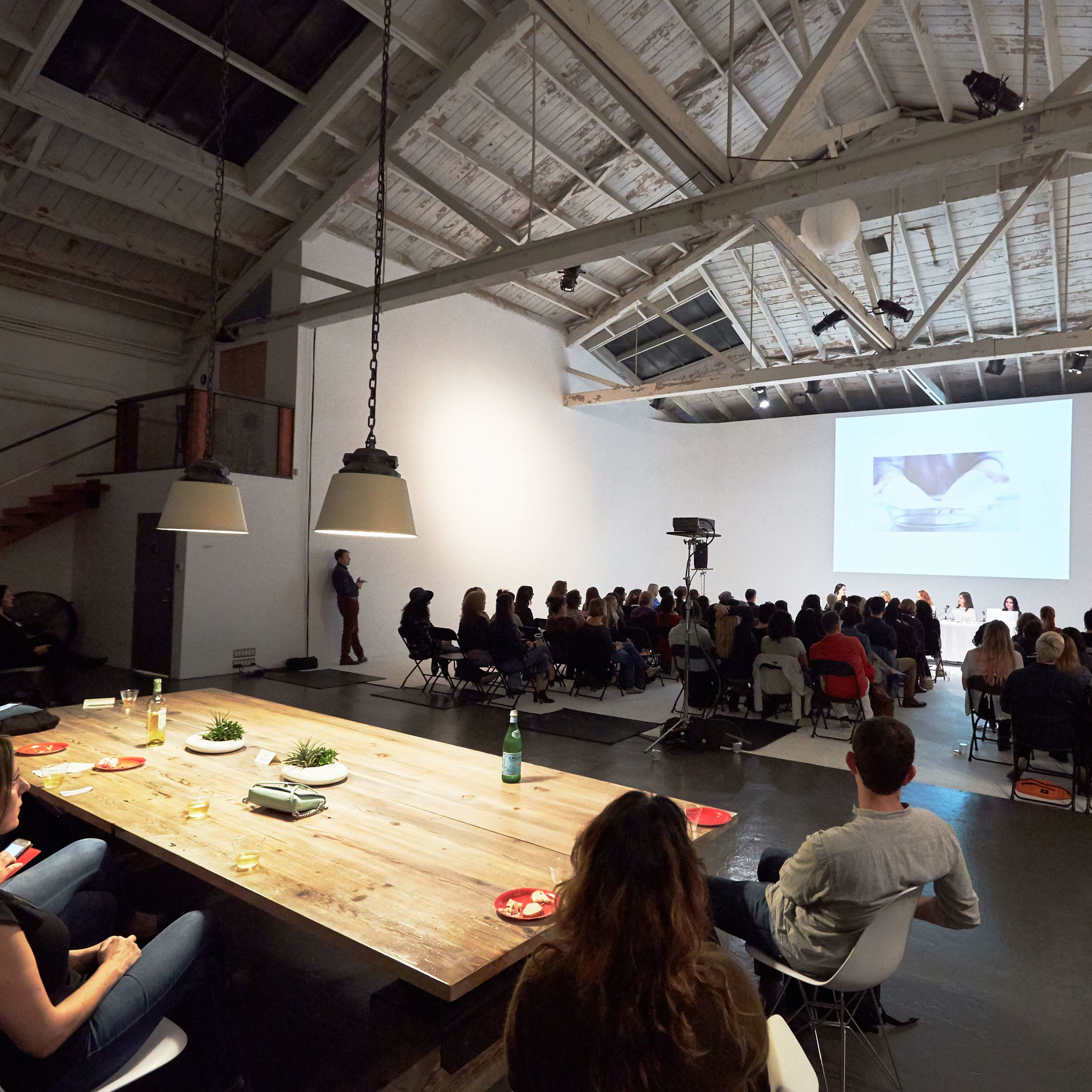 A large audience sits in a modern event space with a presentation screen at the front and a long wooden table in the foreground.