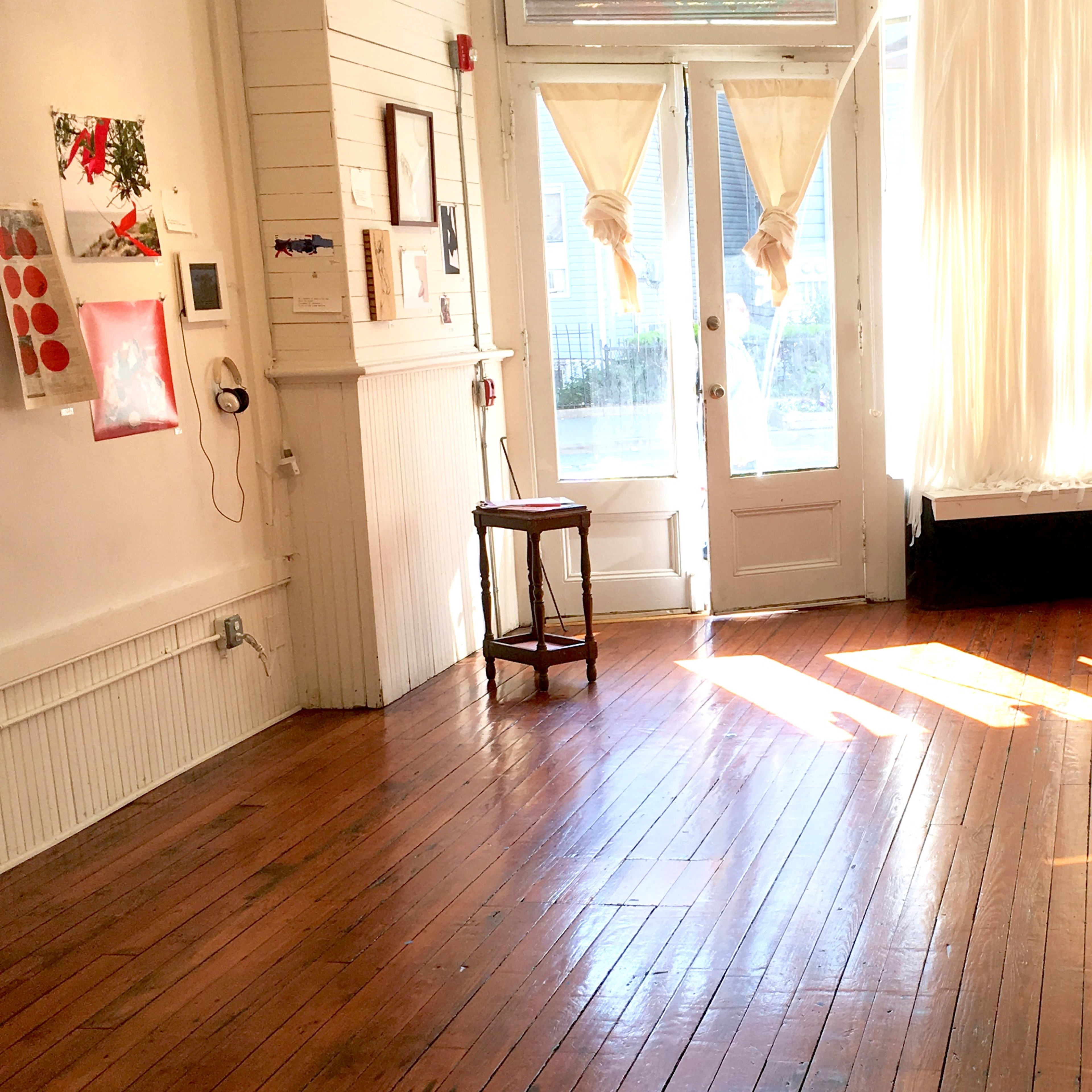 A sunlit room with wooden floors features a chair, large windows with white curtains, and various artworks displayed on the walls.