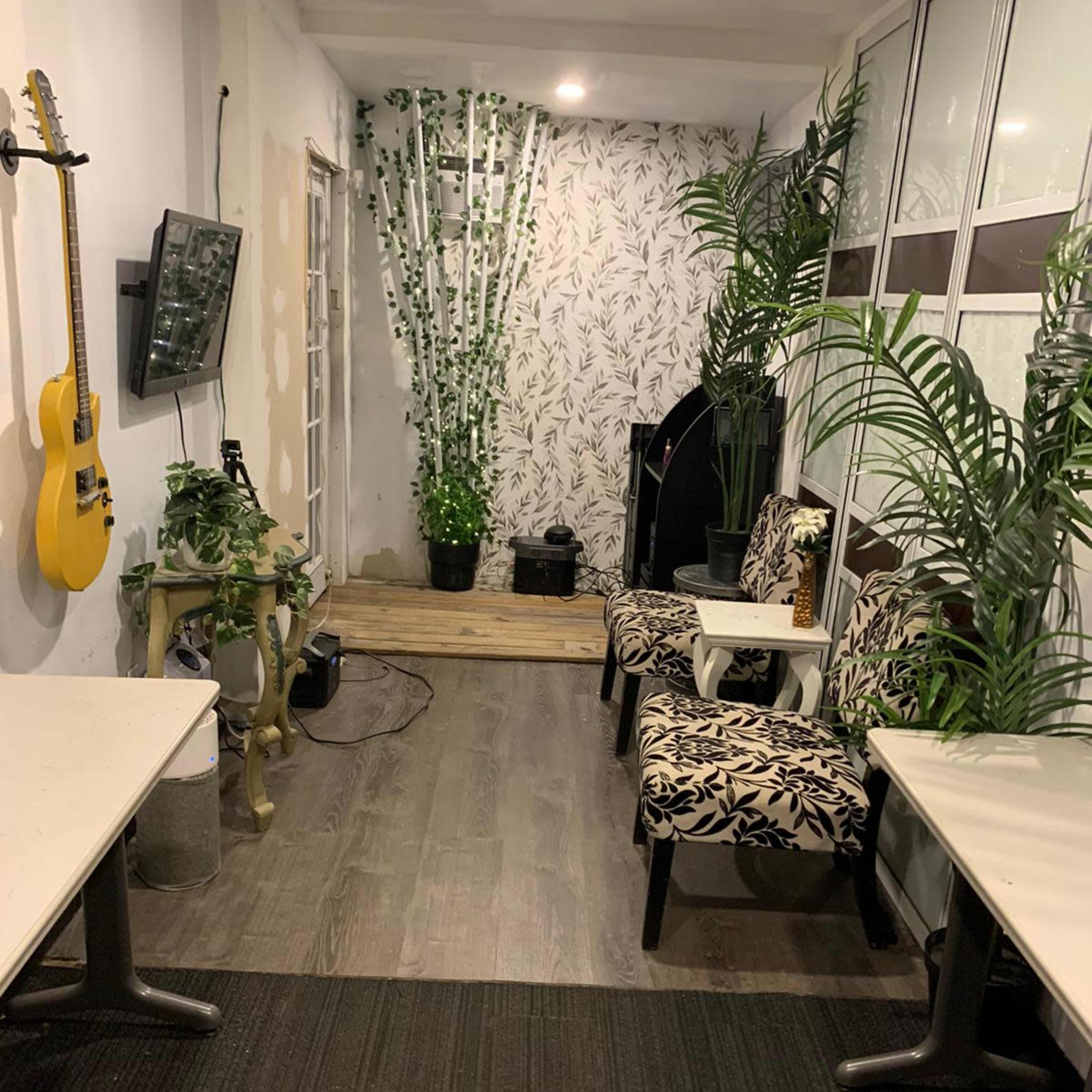 A narrow room with bamboo wallpaper, potted plants, and two patterned chairs, facing a wall-mounted TV and guitar, with wooden flooring and two white tables.