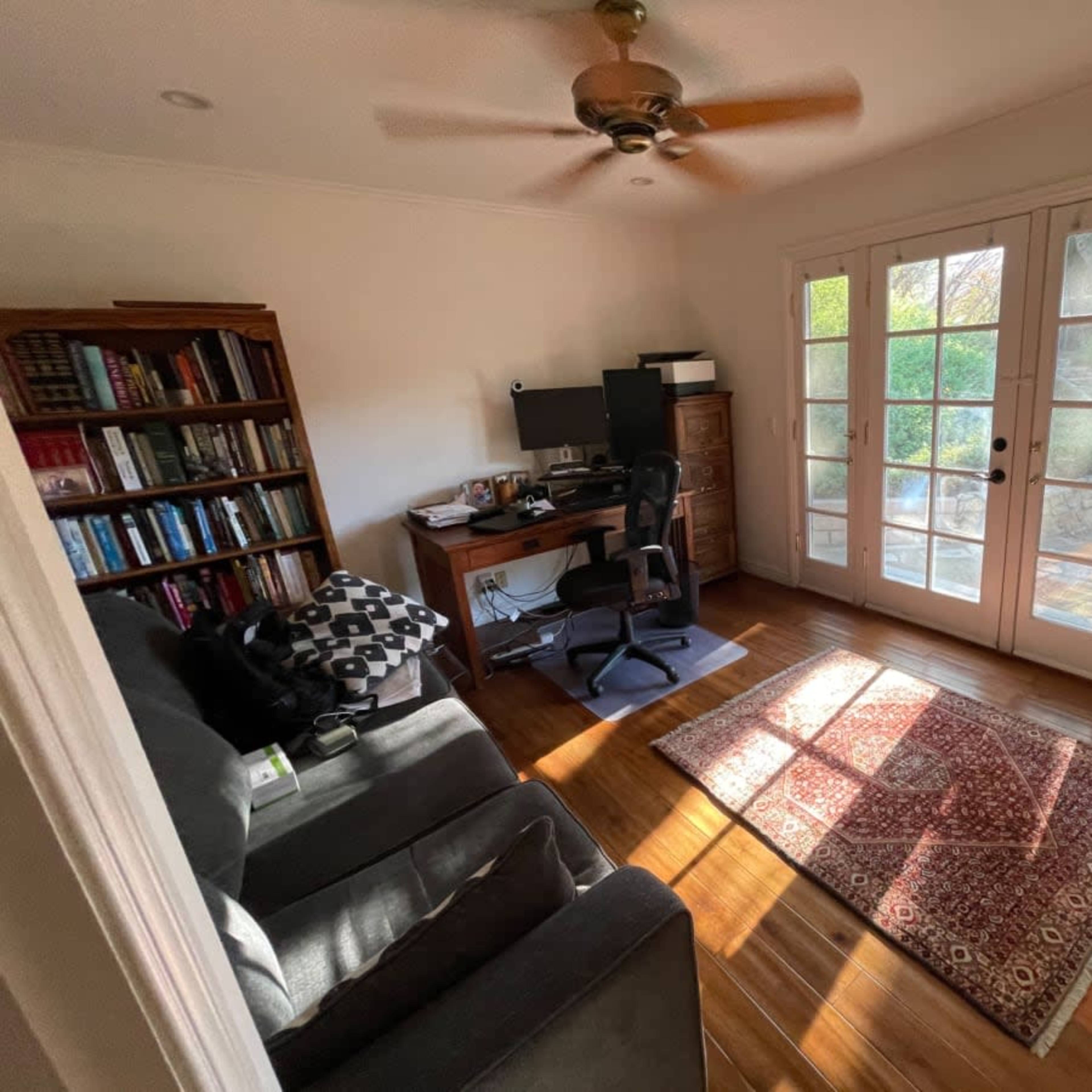 A small home office features a desk with a computer, a chair, a bookshelf, and large French doors that lead to an outdoor area.
