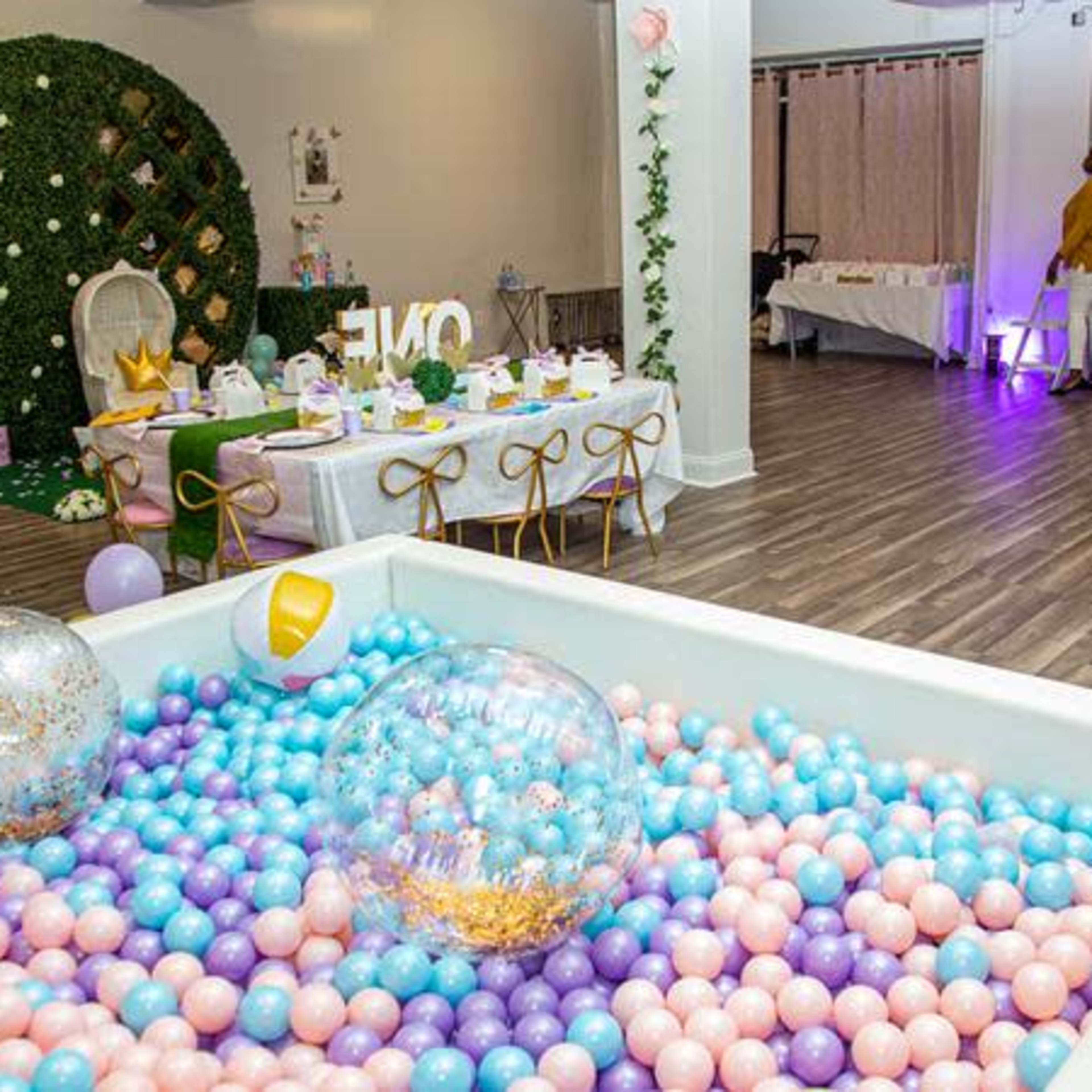 A decorated event space featuring a ball pit filled with pastel-colored balls, tables set for a celebration, and a backdrop with a themed banner.
