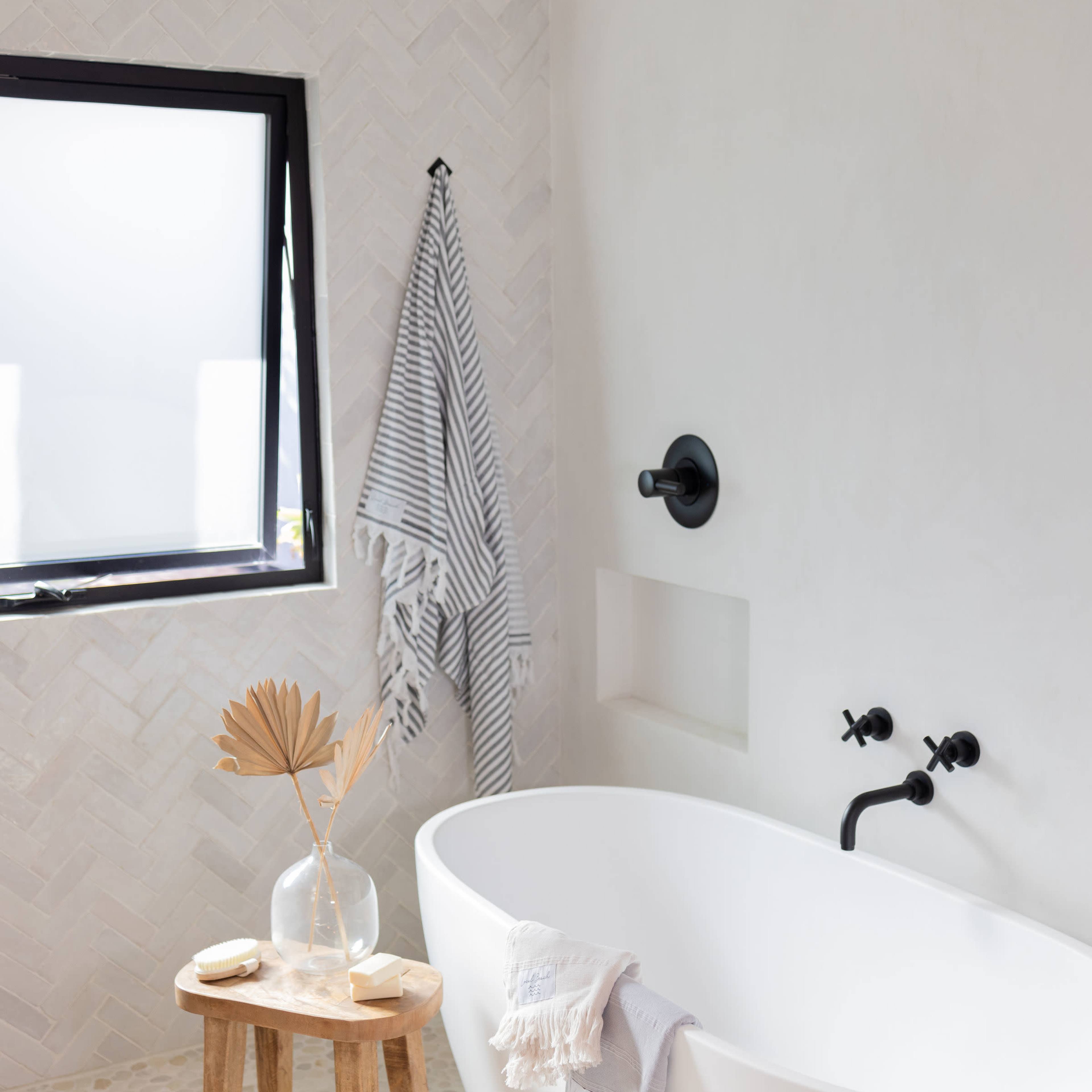 A minimalistic bathroom featuring a freestanding white bathtub, a wooden stool with a decorative vase and dried leaves, and a window with a monochrome striped towel hanging nearby.
