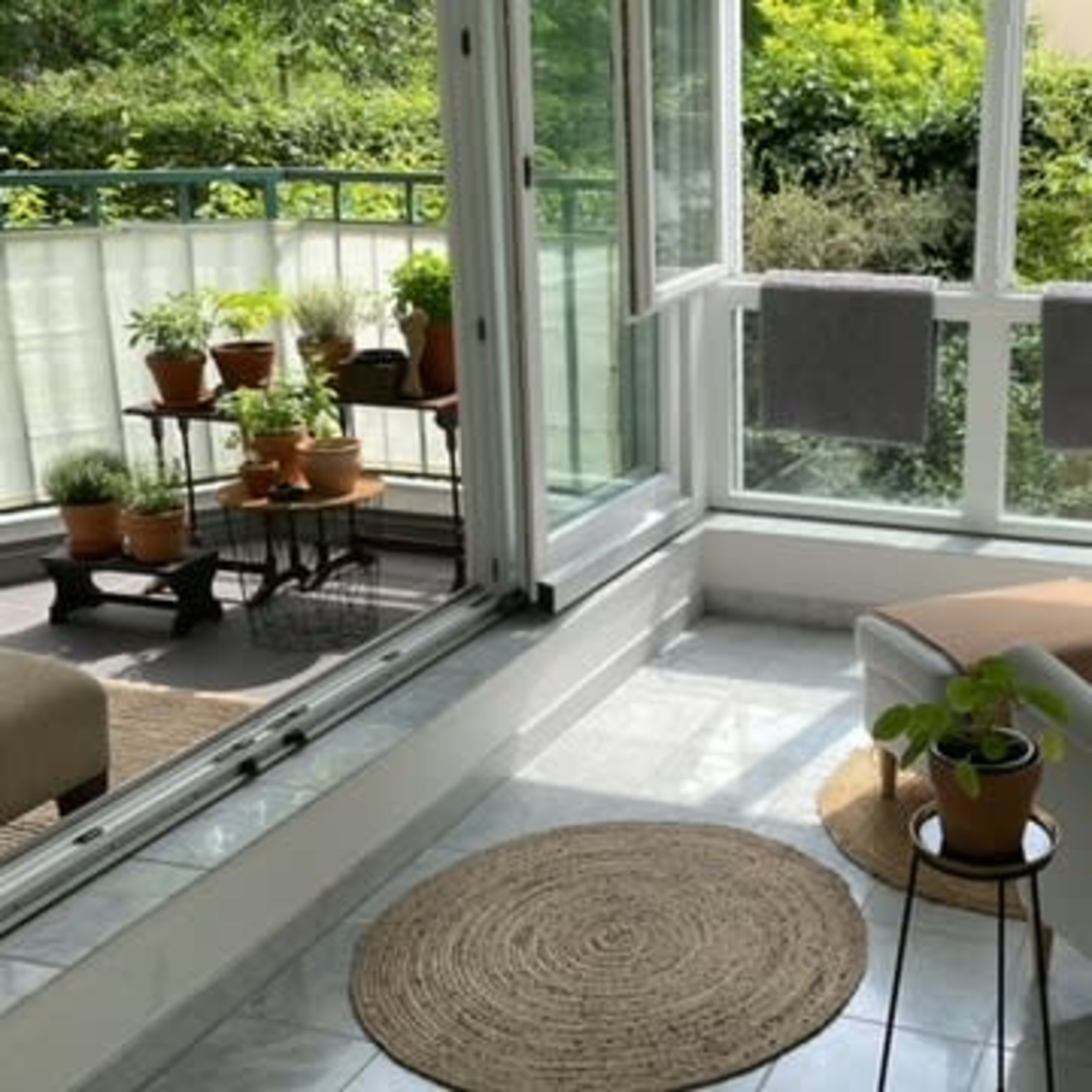 A sunlit sunroom features a cozy chair, a small table with a potted plant, and several plants on a metal cart, surrounded by large windows open to a green view.