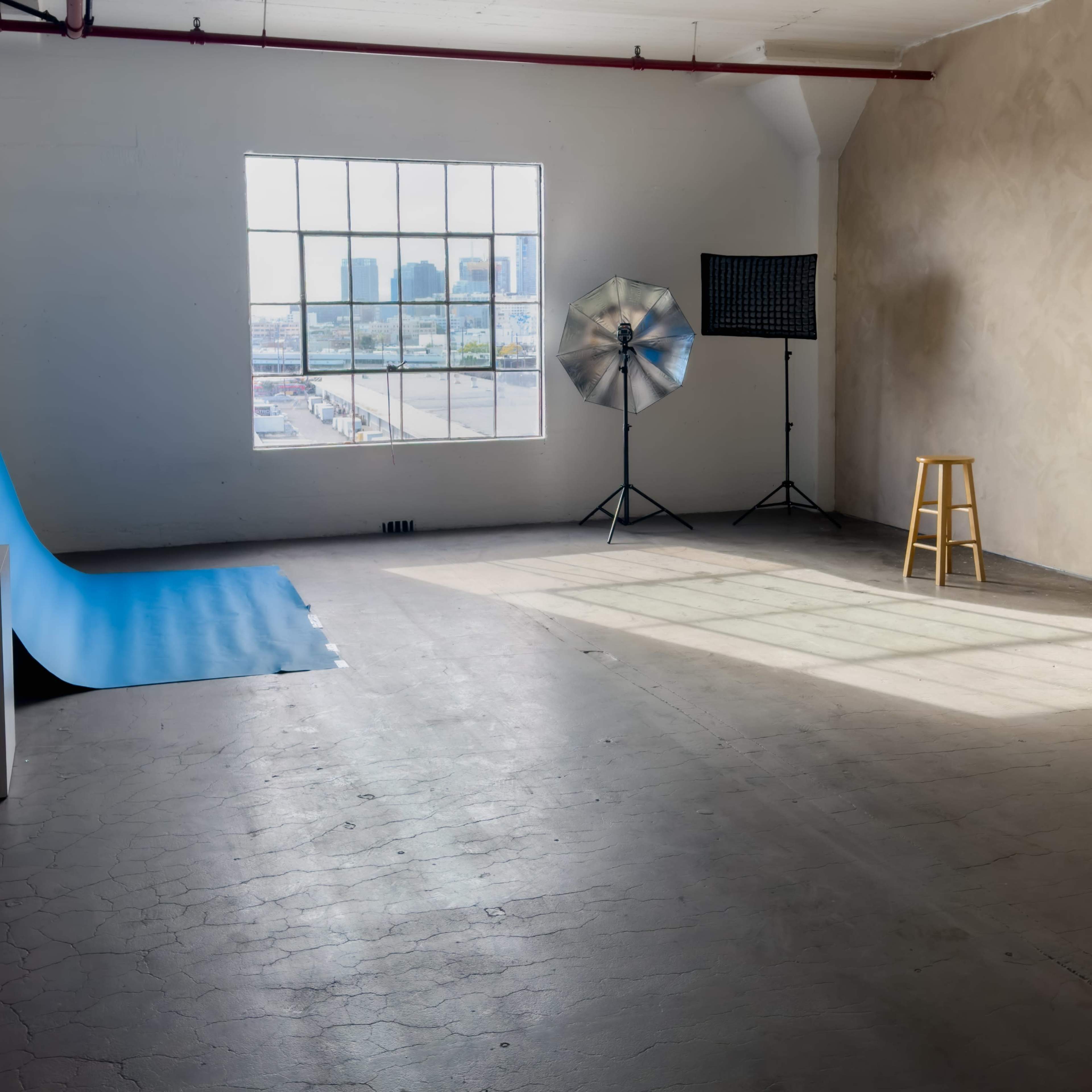 A spacious, well-lit studio with a blue backdrop, a stool, and lighting equipment near a large window.
