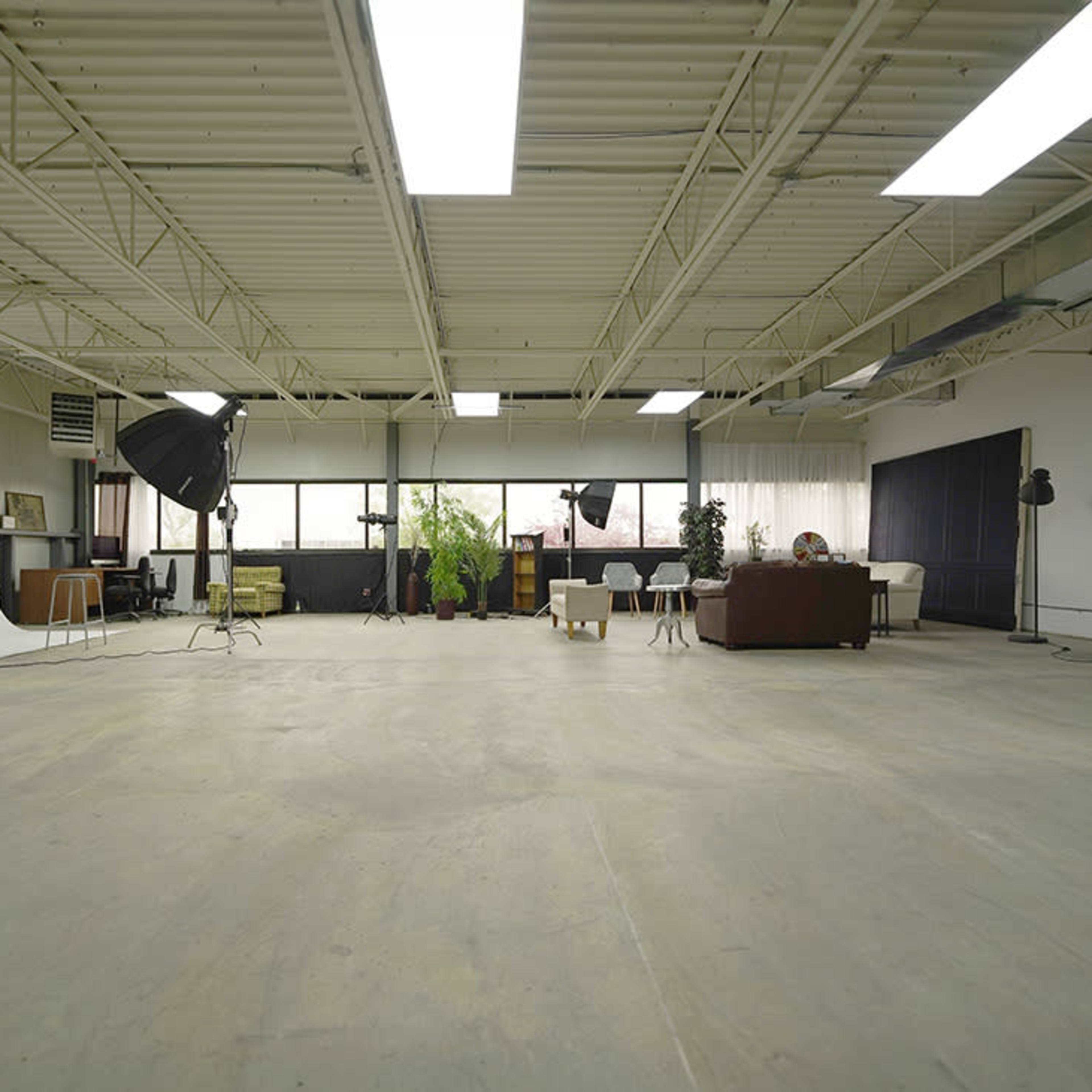A spacious studio with a concrete floor, large windows, and various lighting equipment set up alongside furniture and backdrops.