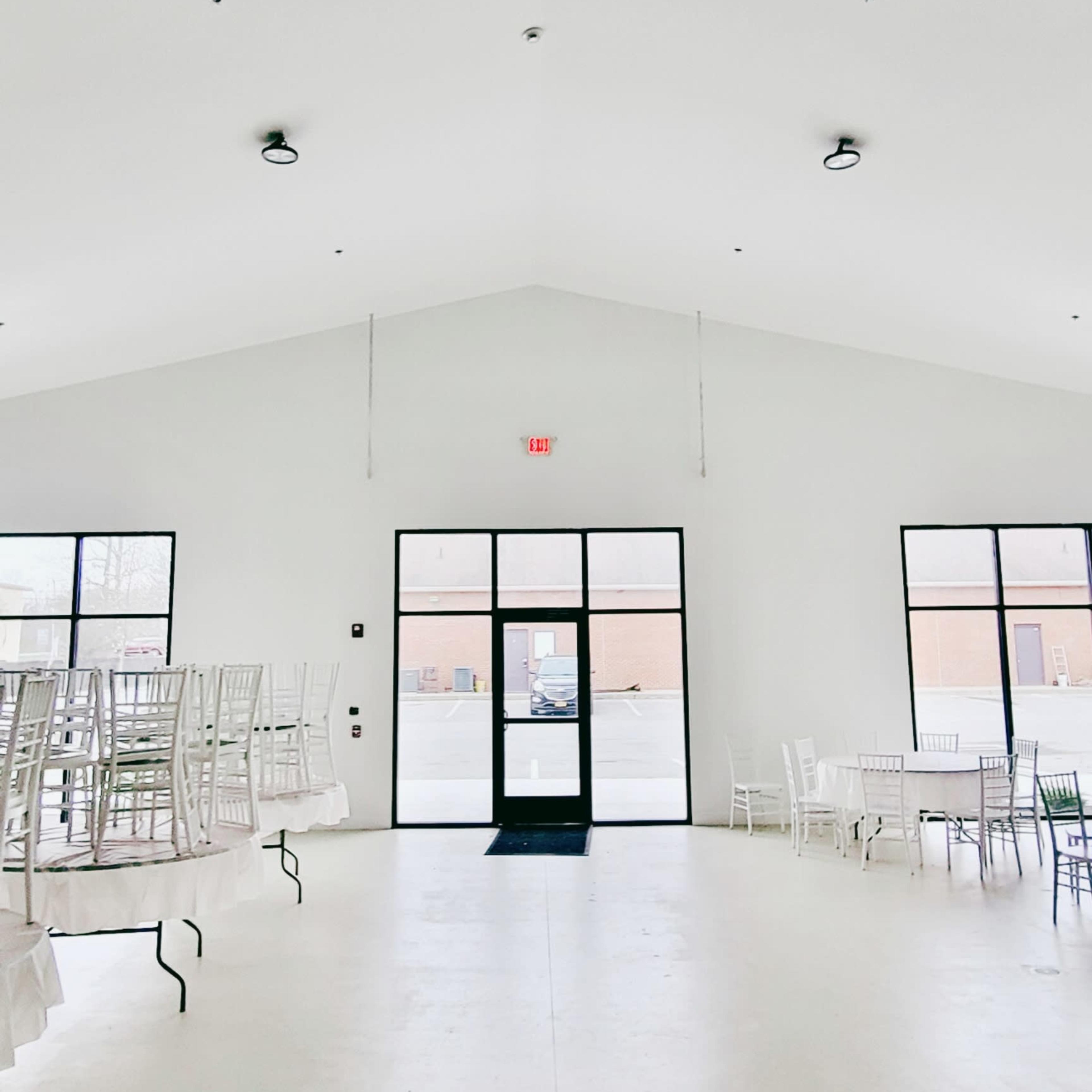 A spacious, empty room with white walls, tall ceilings, and several stacked chairs arranged around the perimeter.