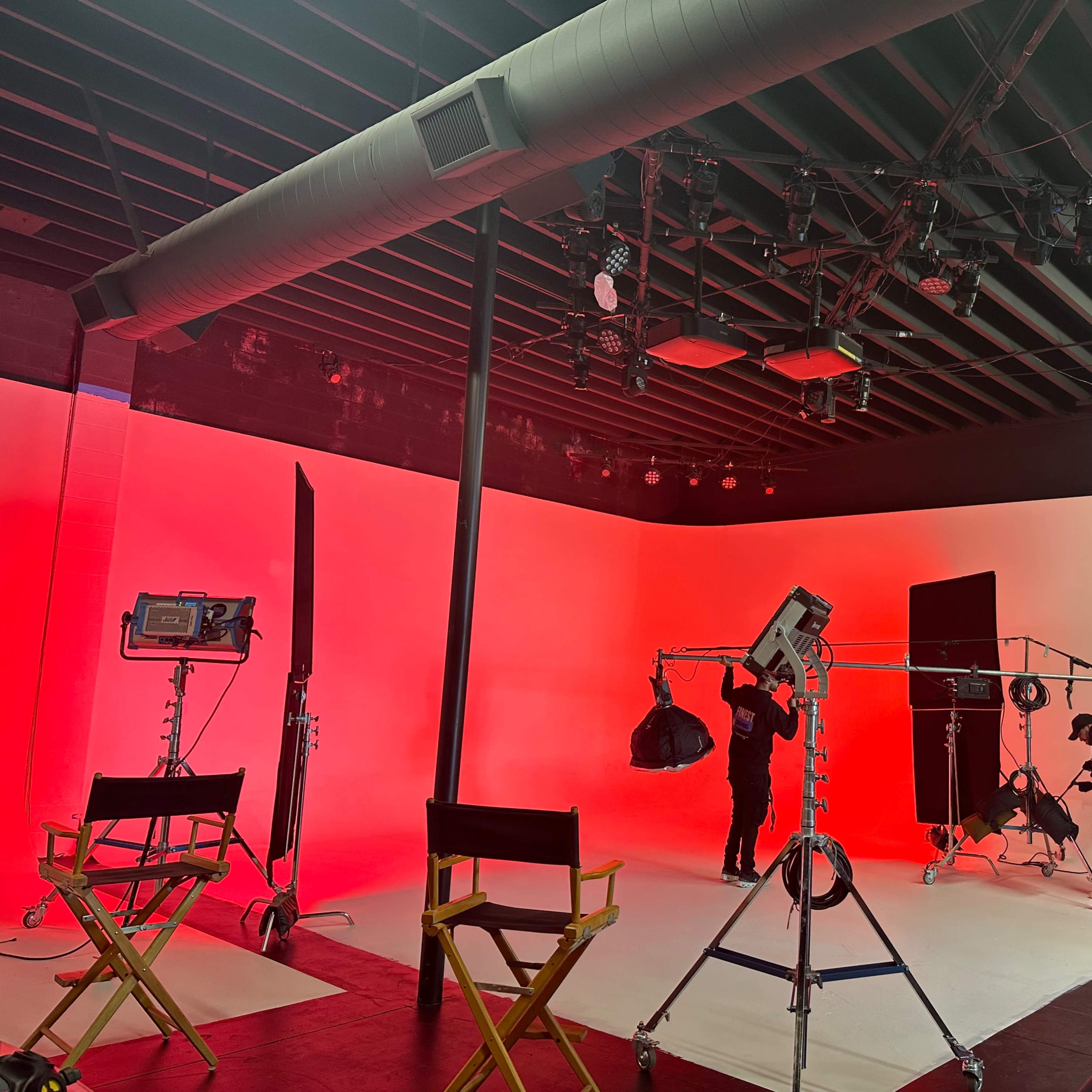 A film studio is set up with multiple lighting rigs and crew members preparing for a shoot against a red and white backdrop.