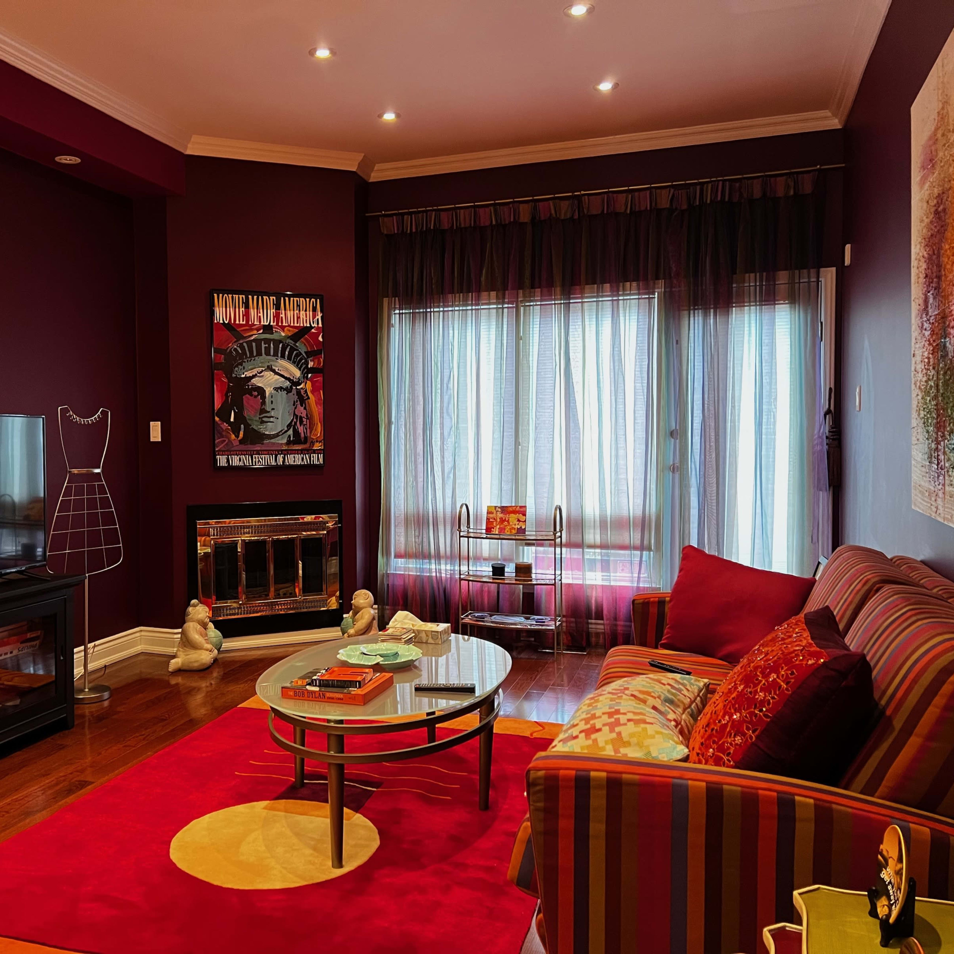 A cozy living room featuring a striped sofa, a round glass coffee table, a television, and a large window covered with sheer curtains.