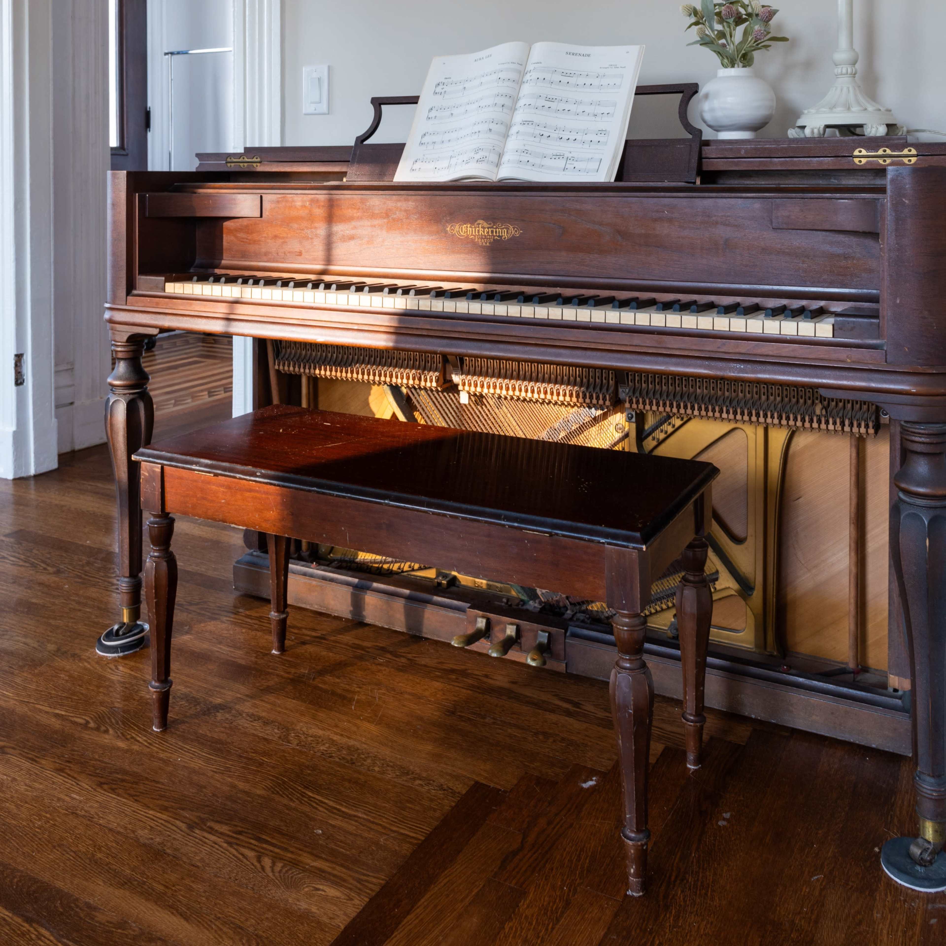 A wooden piano with sheet music on top and a matching bench is positioned in a well-lit room with light-colored walls.