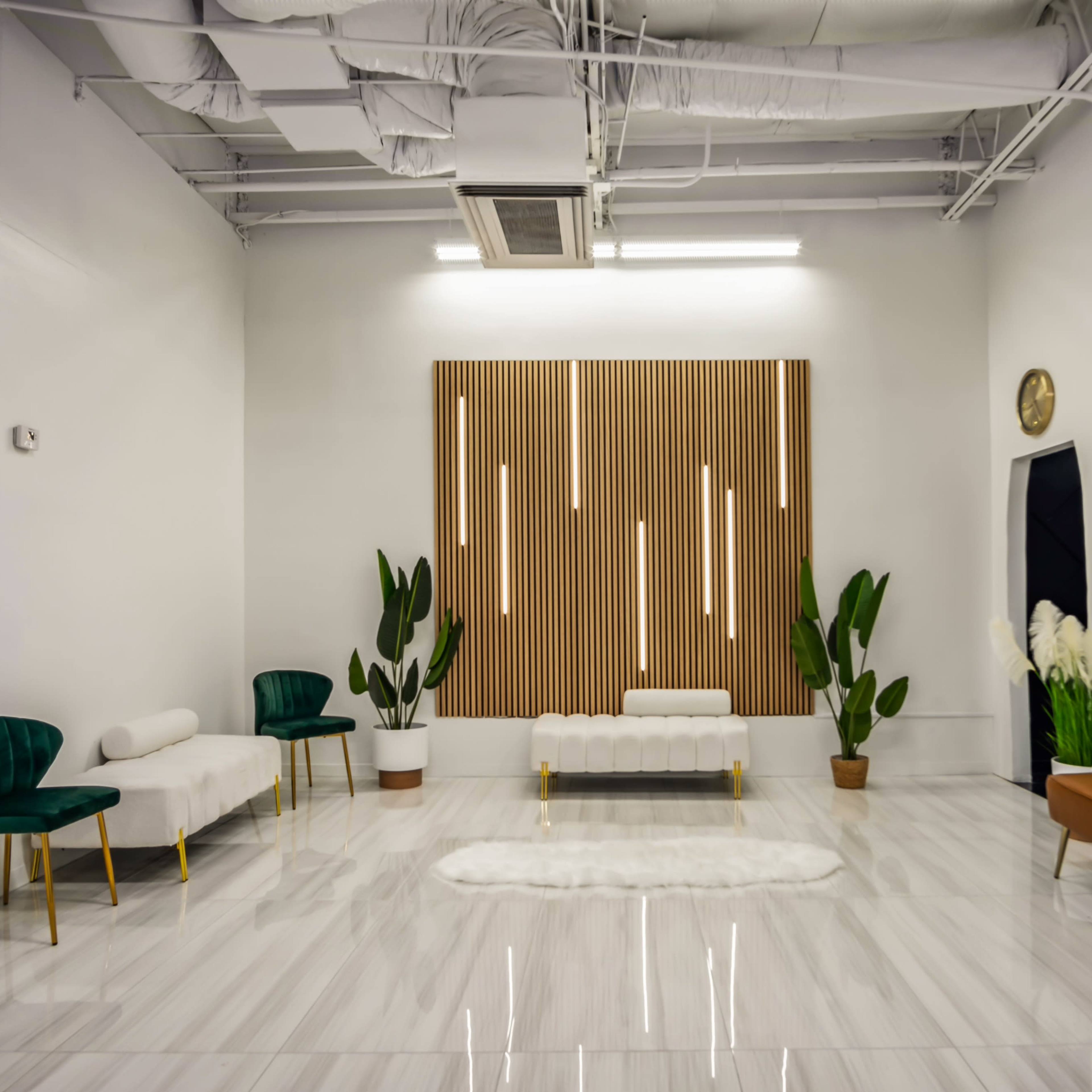 A modern, minimalist interior featuring white walls, a glossy floor, a wooden accent wall with vertical lights, and several pieces of furniture, including two green chairs, a brown sofa, and potted plants.