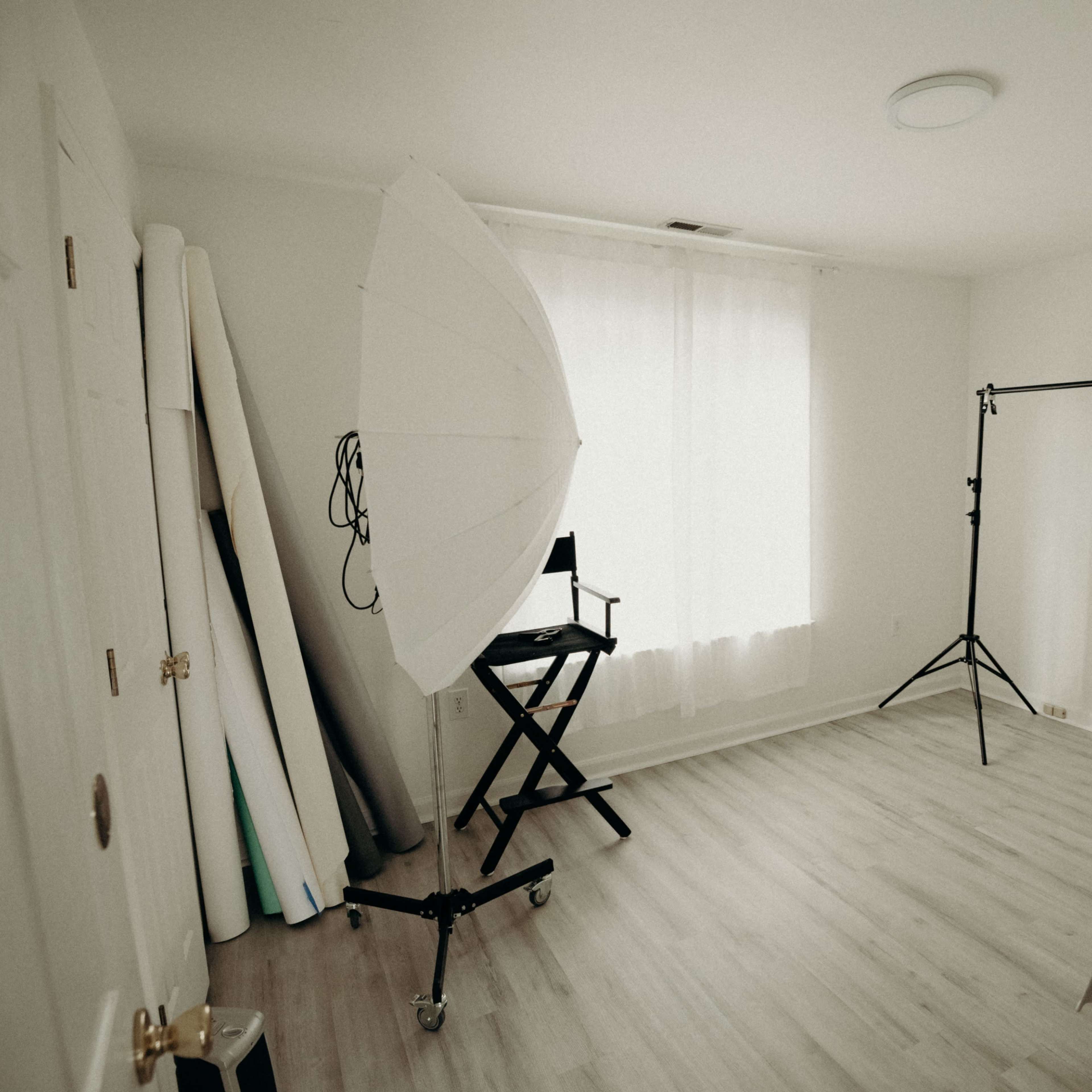 A minimalistic photography studio with a large umbrella light, a portable backdrop stand, and a director's chair in a well-lit room.