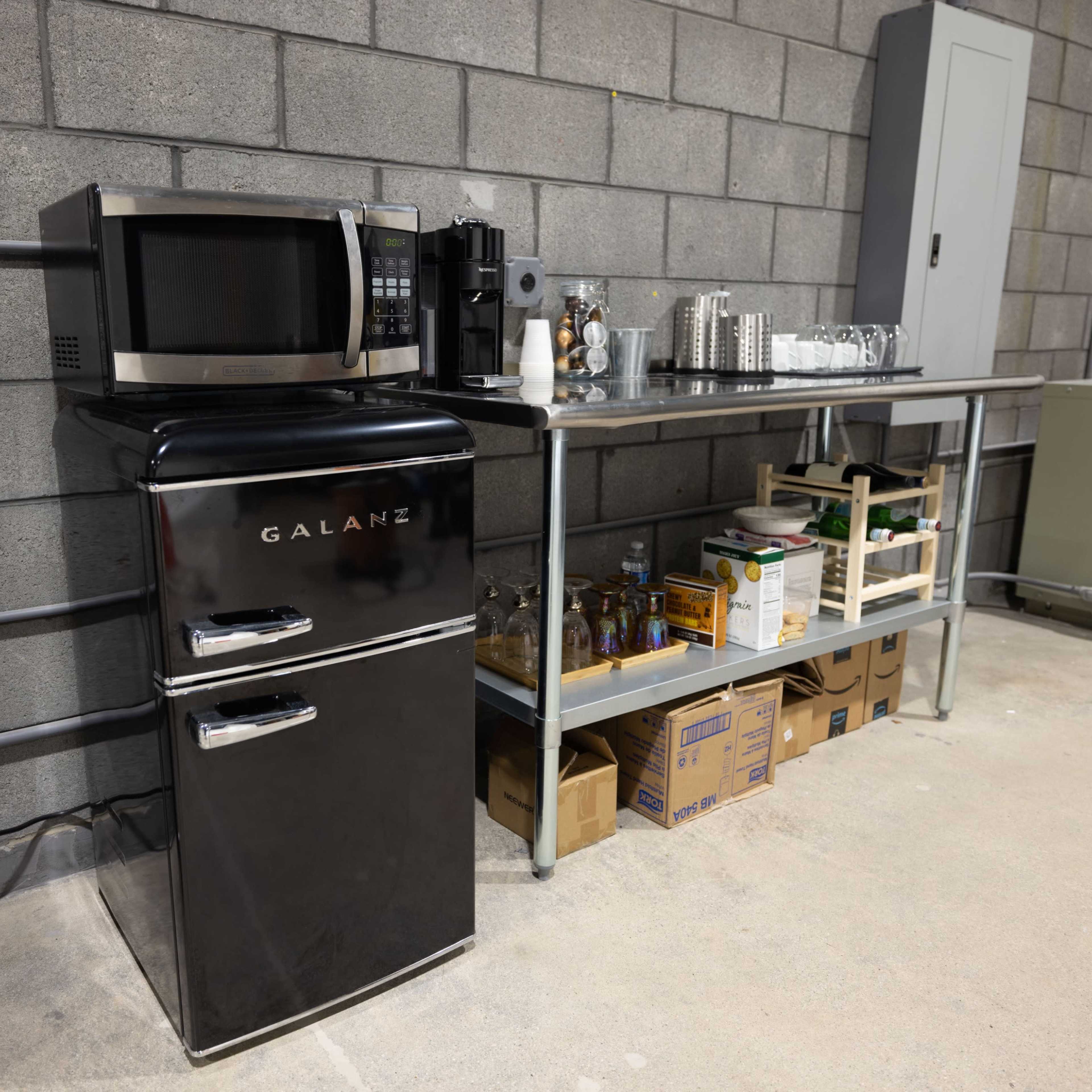 A black Galanz refrigerator and microwave are positioned next to a metal shelving unit filled with various kitchen supplies in a gray-walled space.