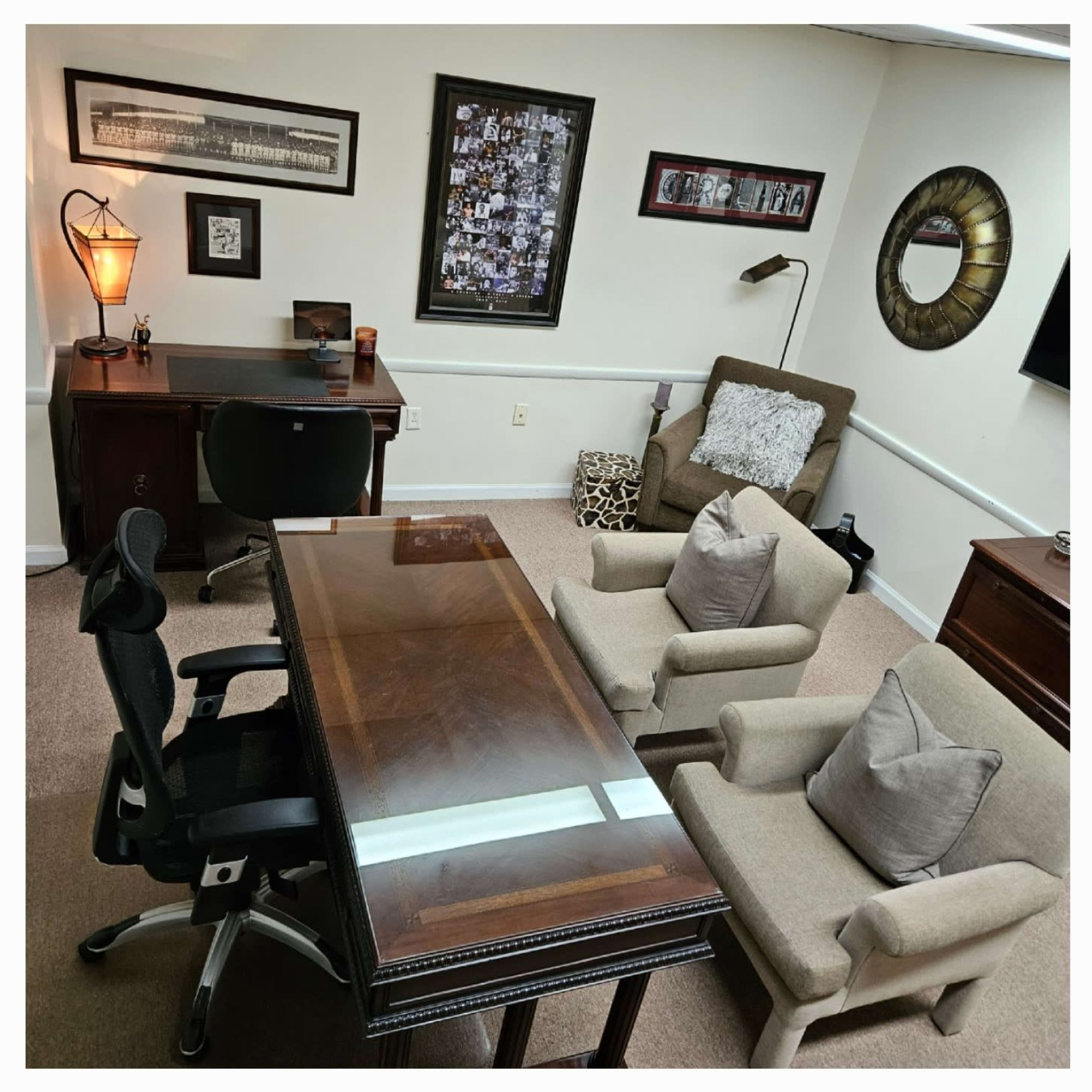 A well-furnished office space featuring a wooden desk, two armchairs, and an office chair, with decorative wall art and a lamp.