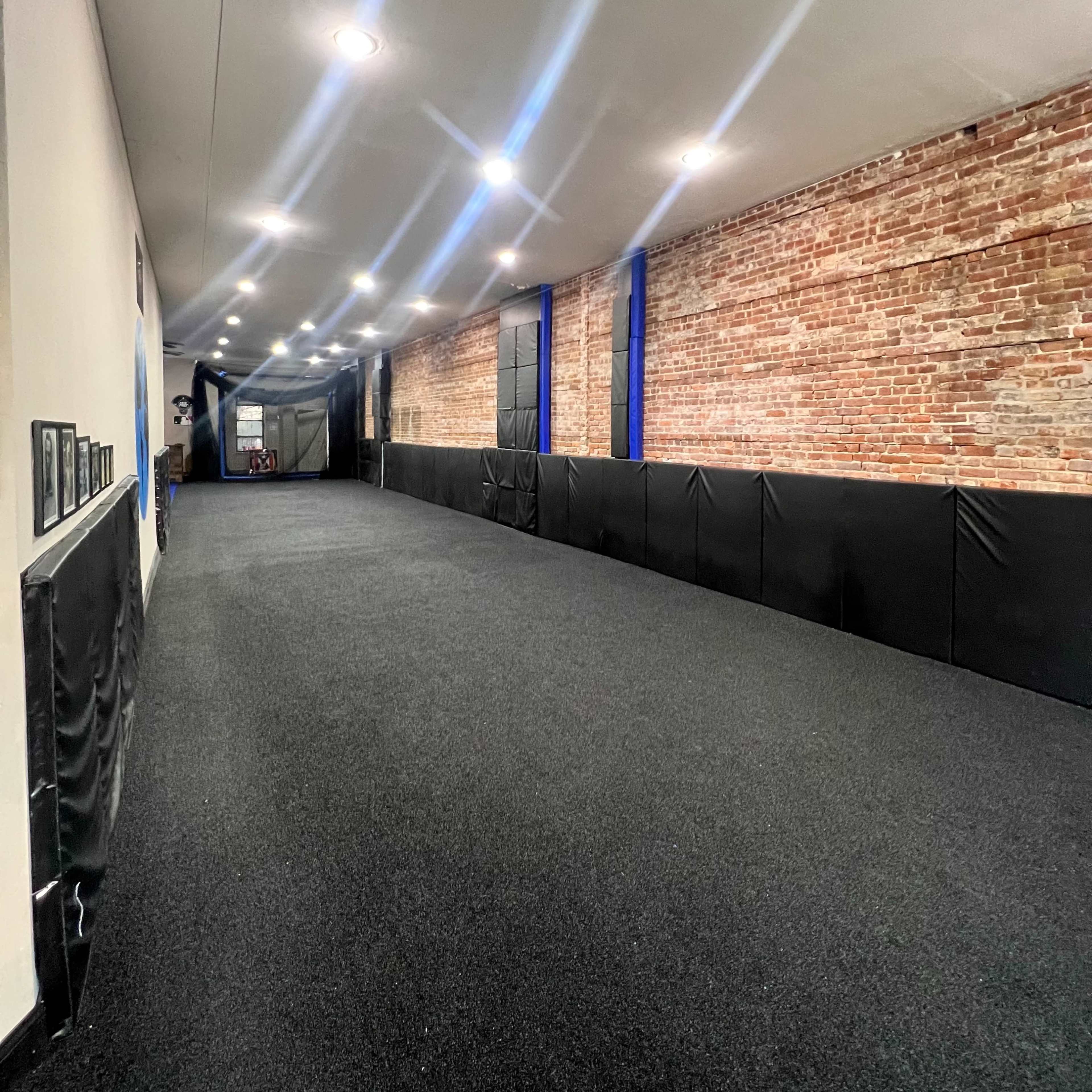 A long, narrow training space with a dark carpeted floor and exposed brick walls, illuminated by overhead lights.