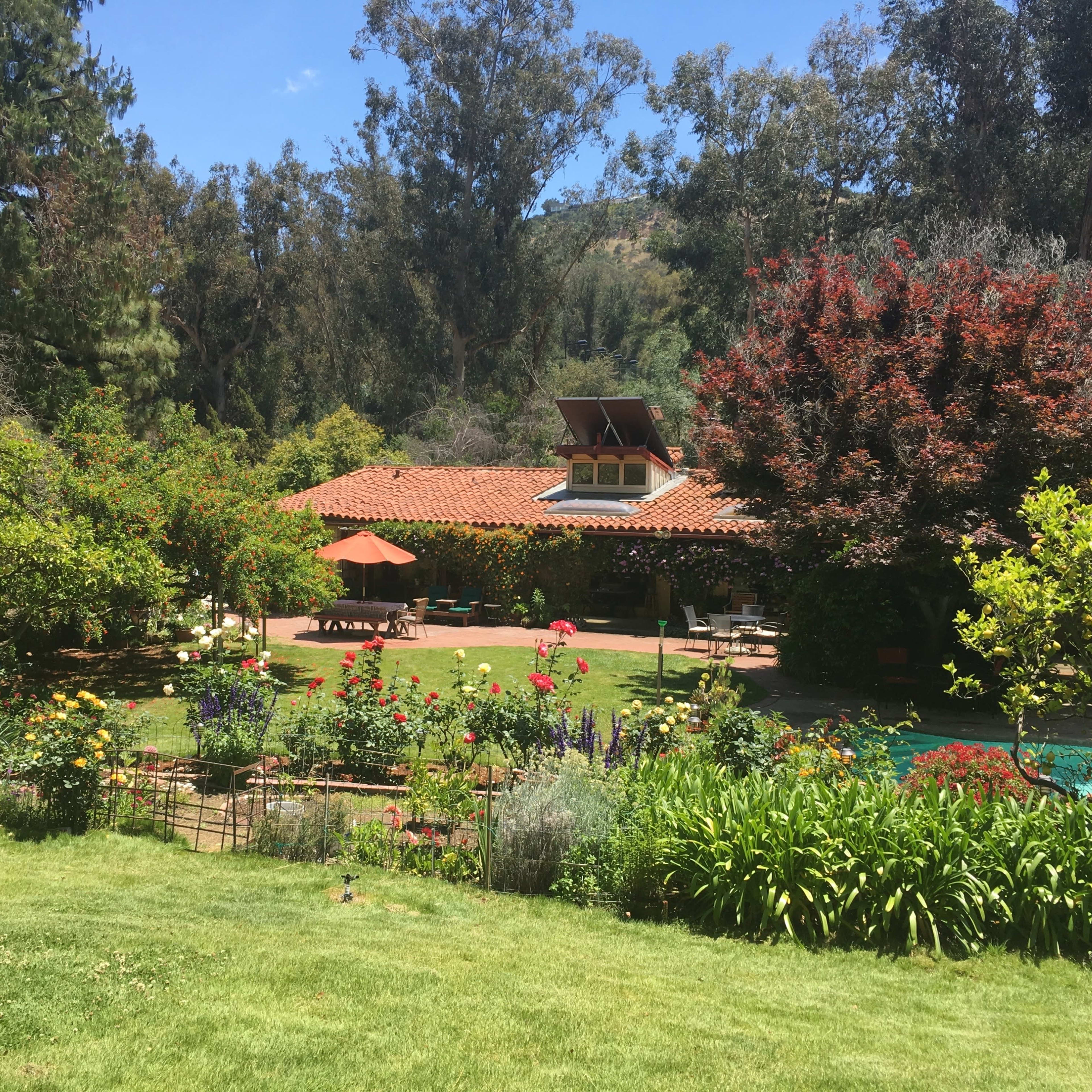 A house with a tiled roof is surrounded by a vibrant garden featuring various flowers, trees, and a swimming pool.