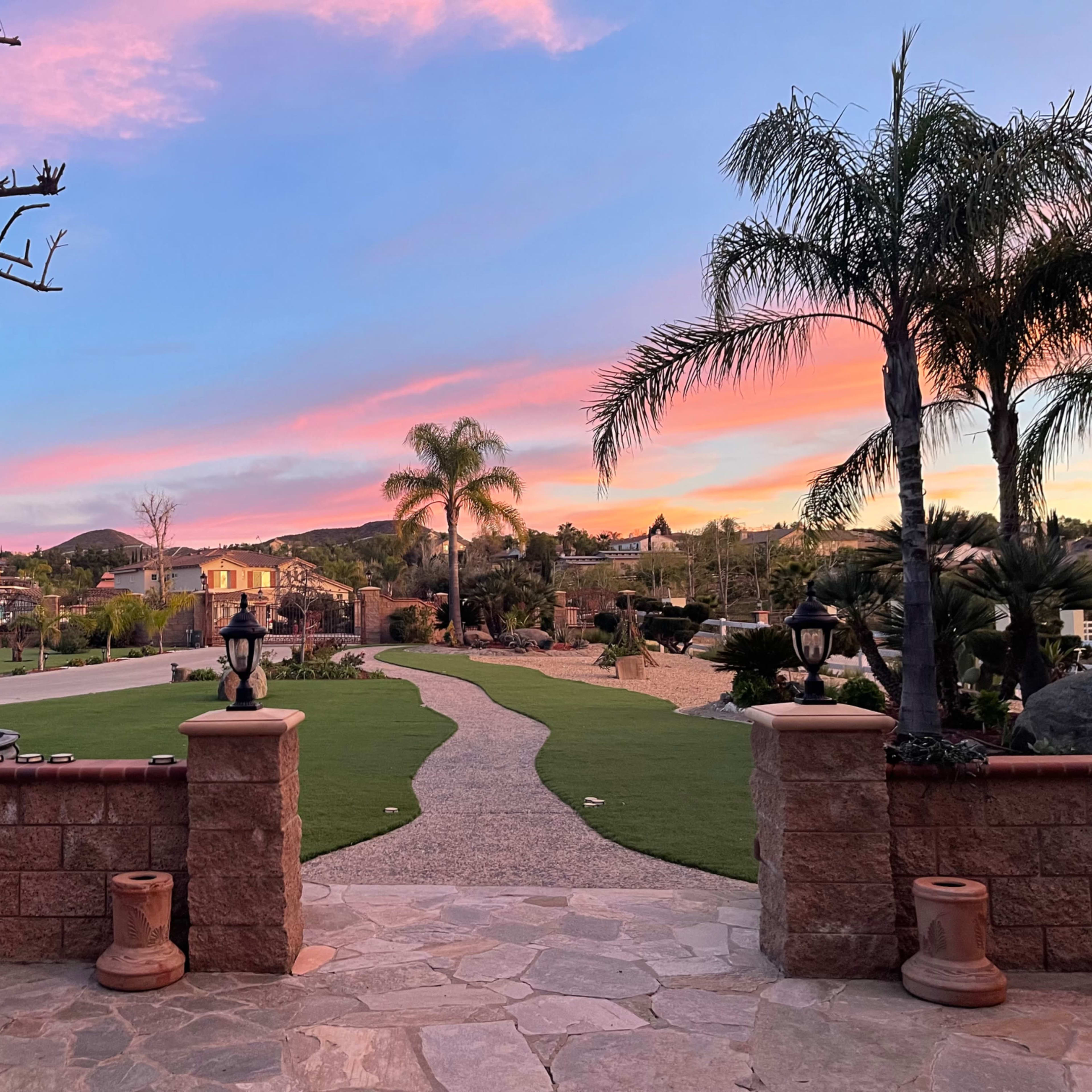 A landscaped garden with a stone pathway leading towards a fountain, framed by palm trees and a colorful sunset sky.