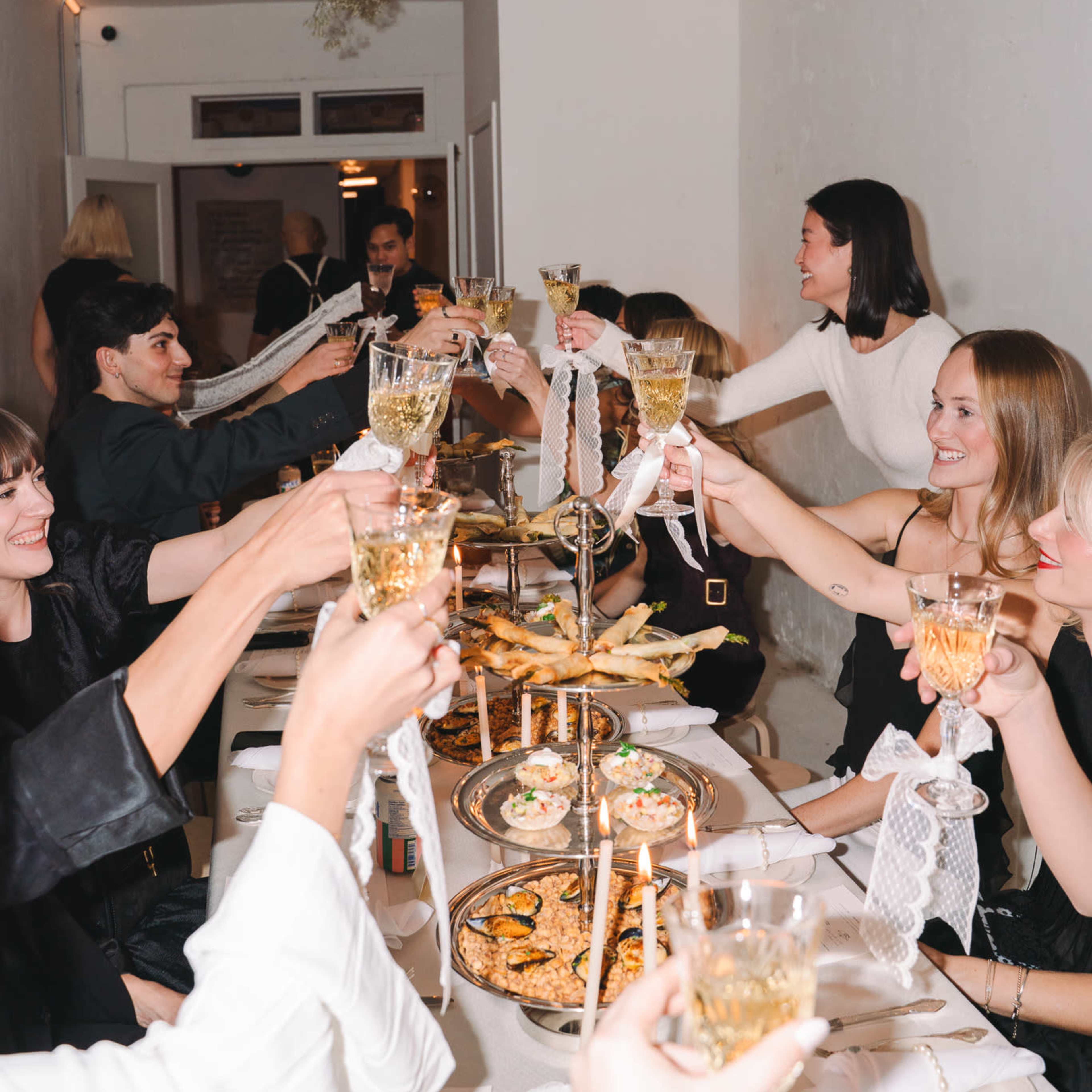 A group of people at a long table raise their glasses in a toast, surrounded by various dishes and decorations in a dimly lit setting.