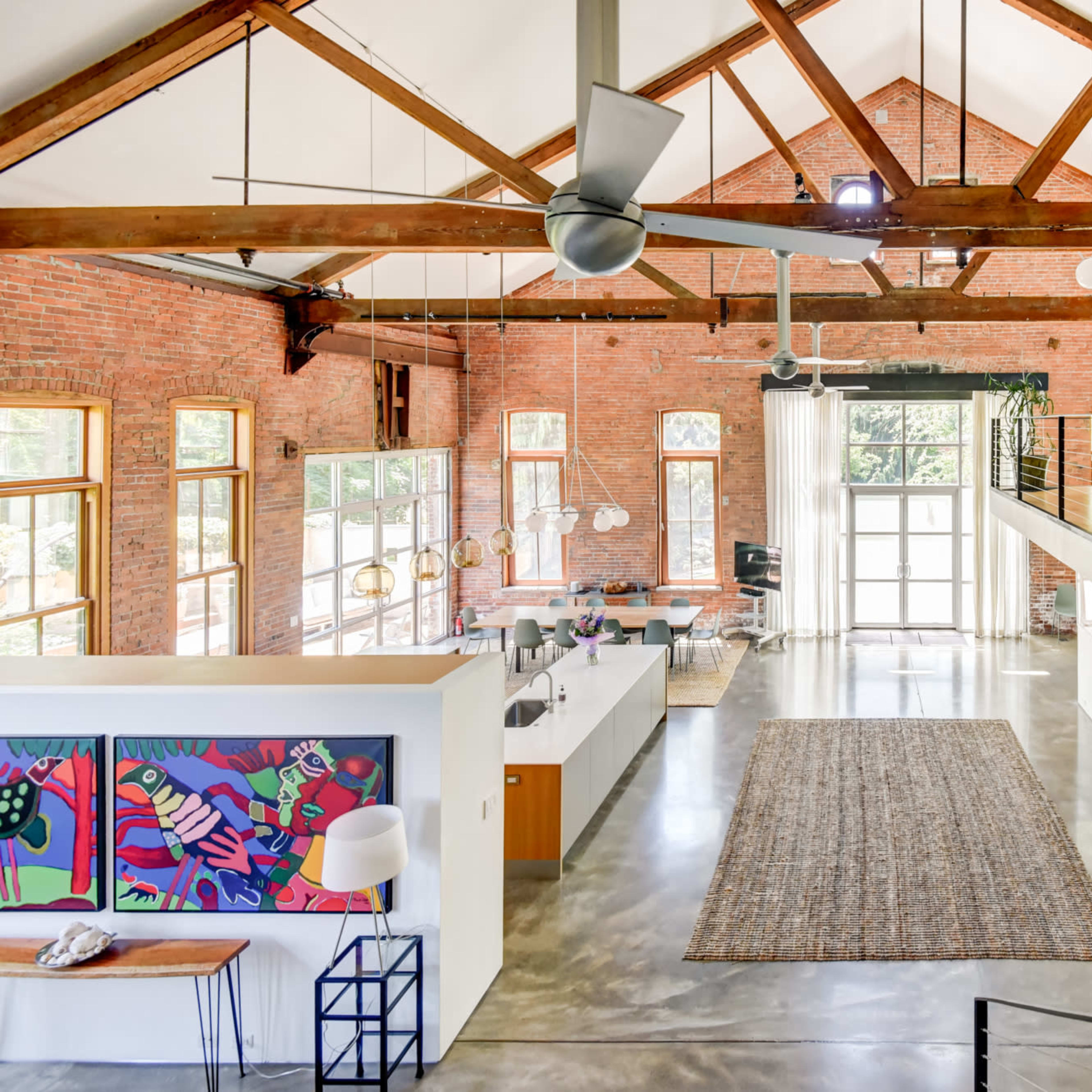 A spacious, modern interior with exposed brick walls, high wood-beamed ceilings, and large windows, featuring an open layout connecting the kitchen and living areas.