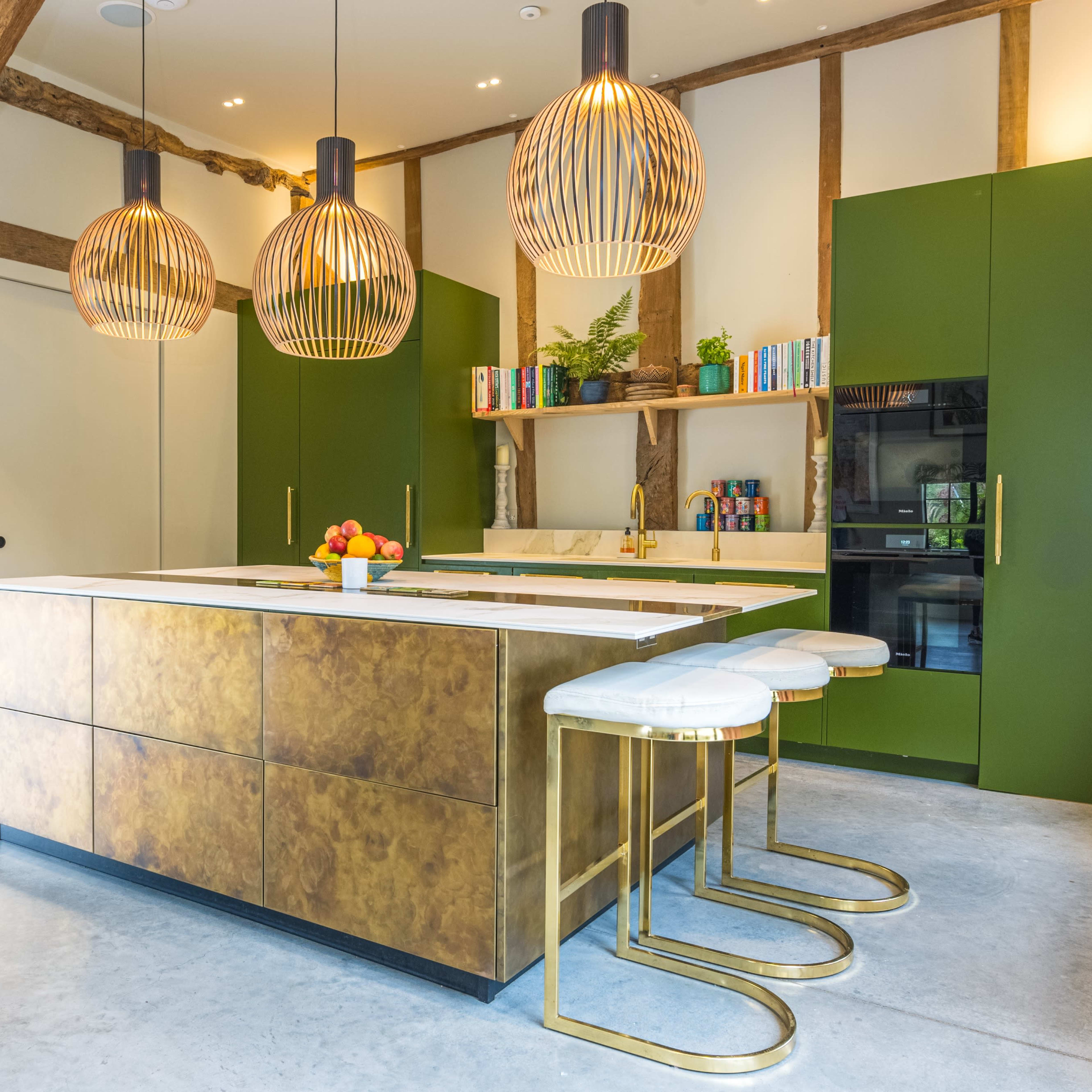 A modern kitchen features a central island with a gold finish, three pendant lights, and green cabinetry, set against exposed wooden beams.