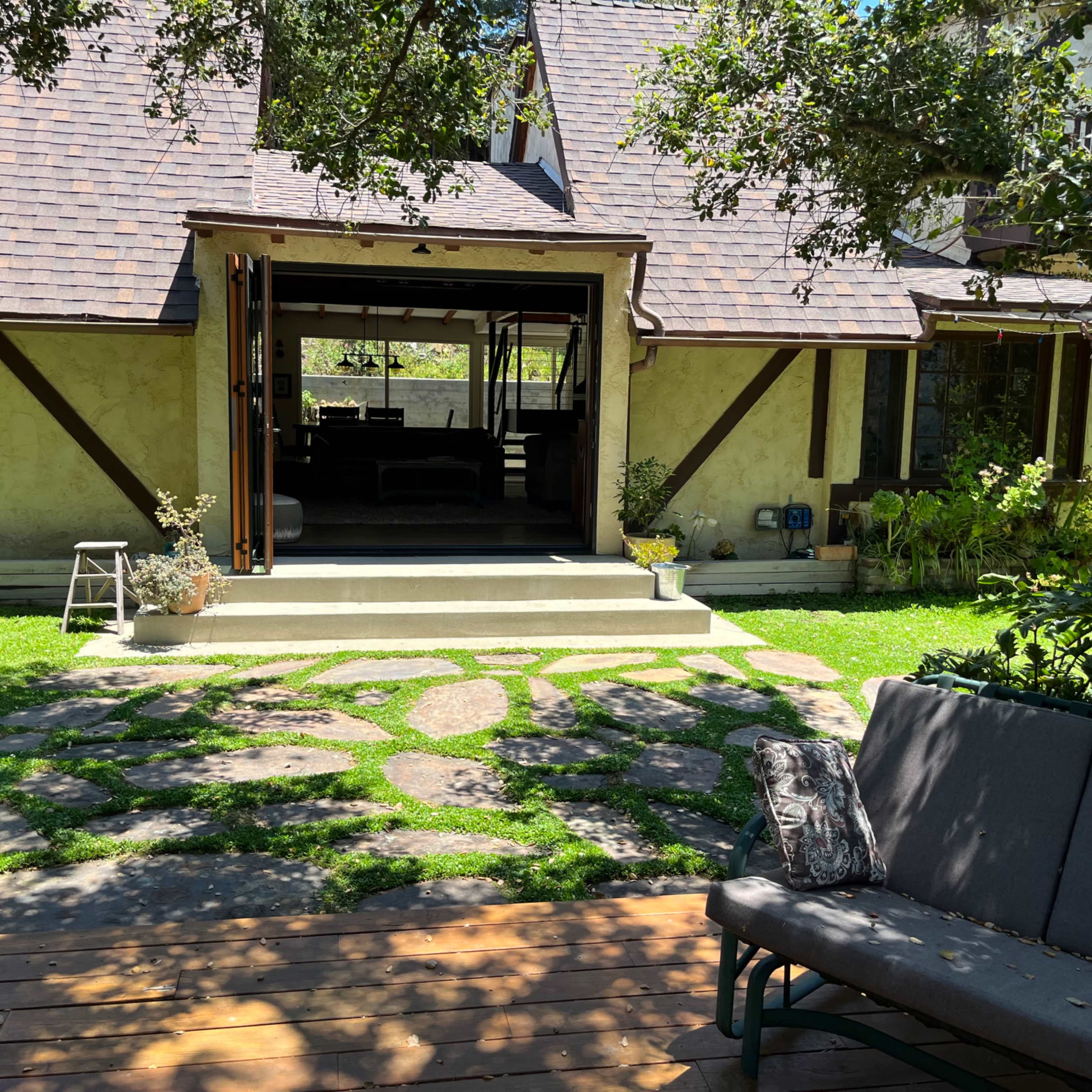 A backyard with a stone pathway leading to a house with a sloped roof and open doors, surrounded by greenery and outdoor seating.